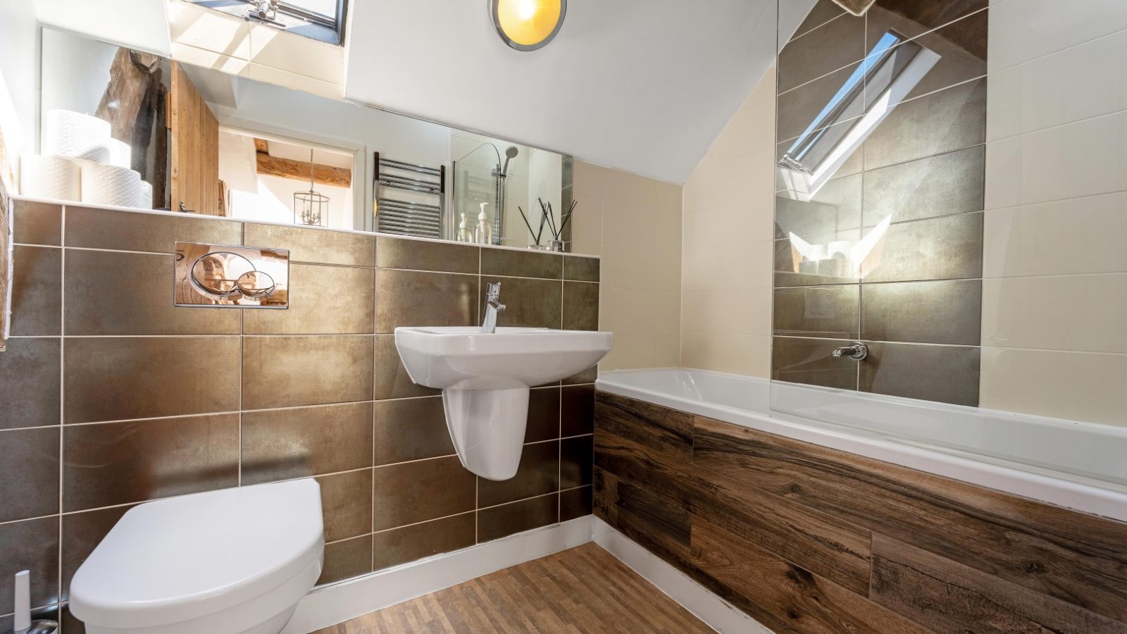 A modern bathroom at The Water Mill features a wall-mounted toilet, pedestal sink, tub with wooden paneling, and large brown tiles.