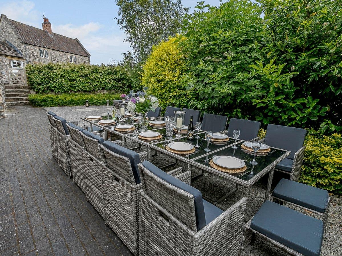 Outdoor dining table set for twelve with wicker chairs and a glass top, nestled by The Water Mill near a stone house.