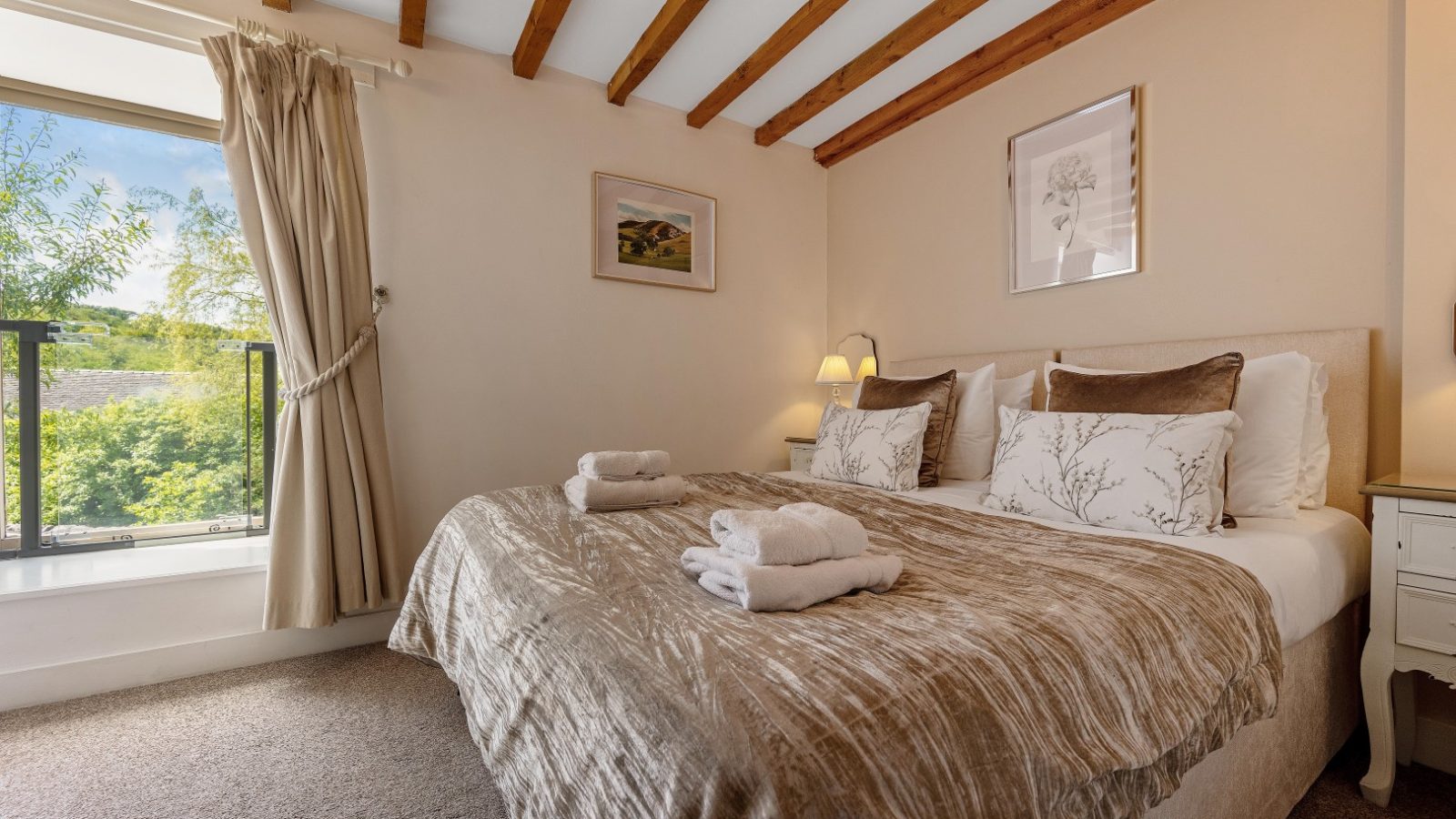 A cozy bedroom at The Water Mill, with a double bed, folded towels, decorative pillows, and window views of greenery.