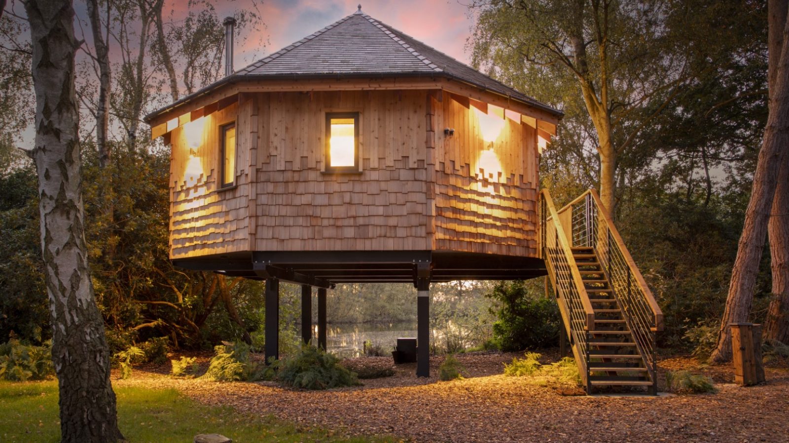 Treetop Hideaway: A wooden treehouse with lit windows on stilts, surrounded by trees at dusk, accessible by staircase.