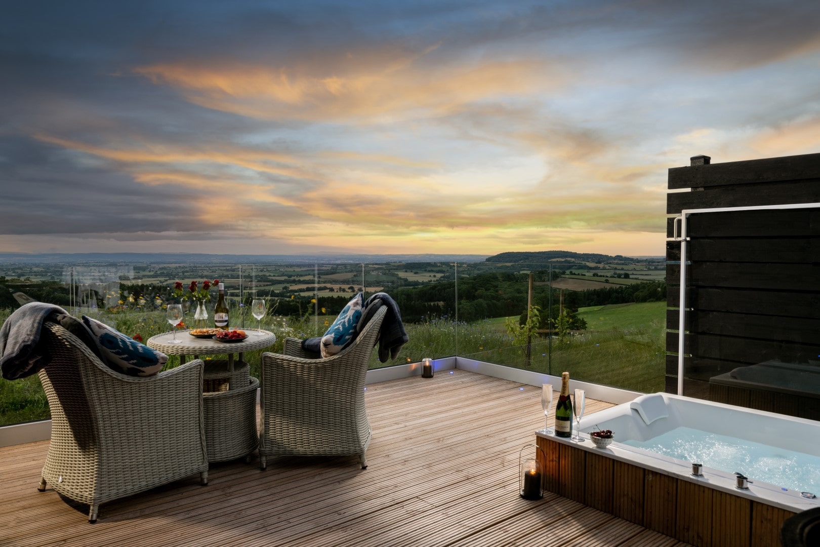 A romantic dog friendly break break at a holiday home with a deck with wicker chairs, table with wine and snacks, and jacuzzi overlooking a scenic sunset.