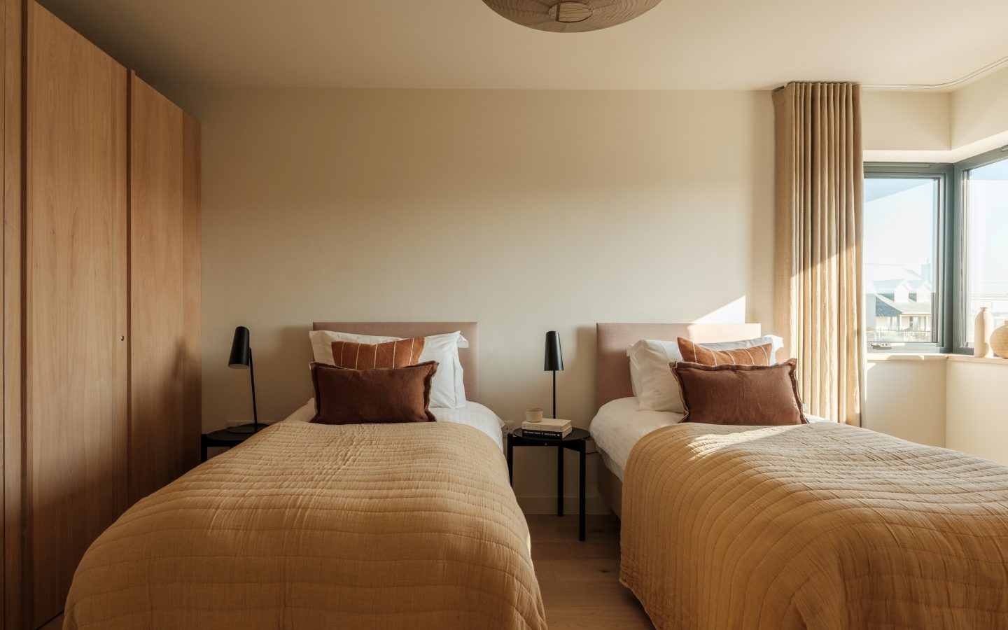 A minimalist bedroom with twin beds, beige bedding, Anterre wardrobe, and a large window. Natural light fills the room.