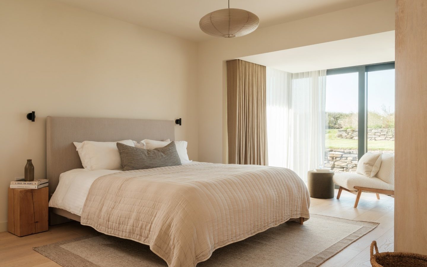 A modern Anterre bedroom in a beige palette, with bed, nightstand, armchair, and a window adorned with sheer curtains.