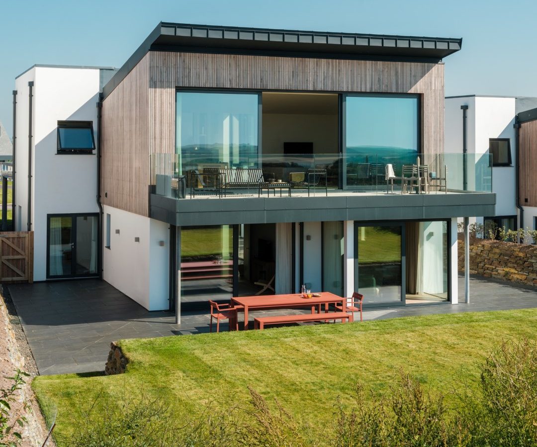 Modern two-story house in Highcliffe, featuring large glass windows and balcony, with a yard and wooden picnic table on a sunny day.