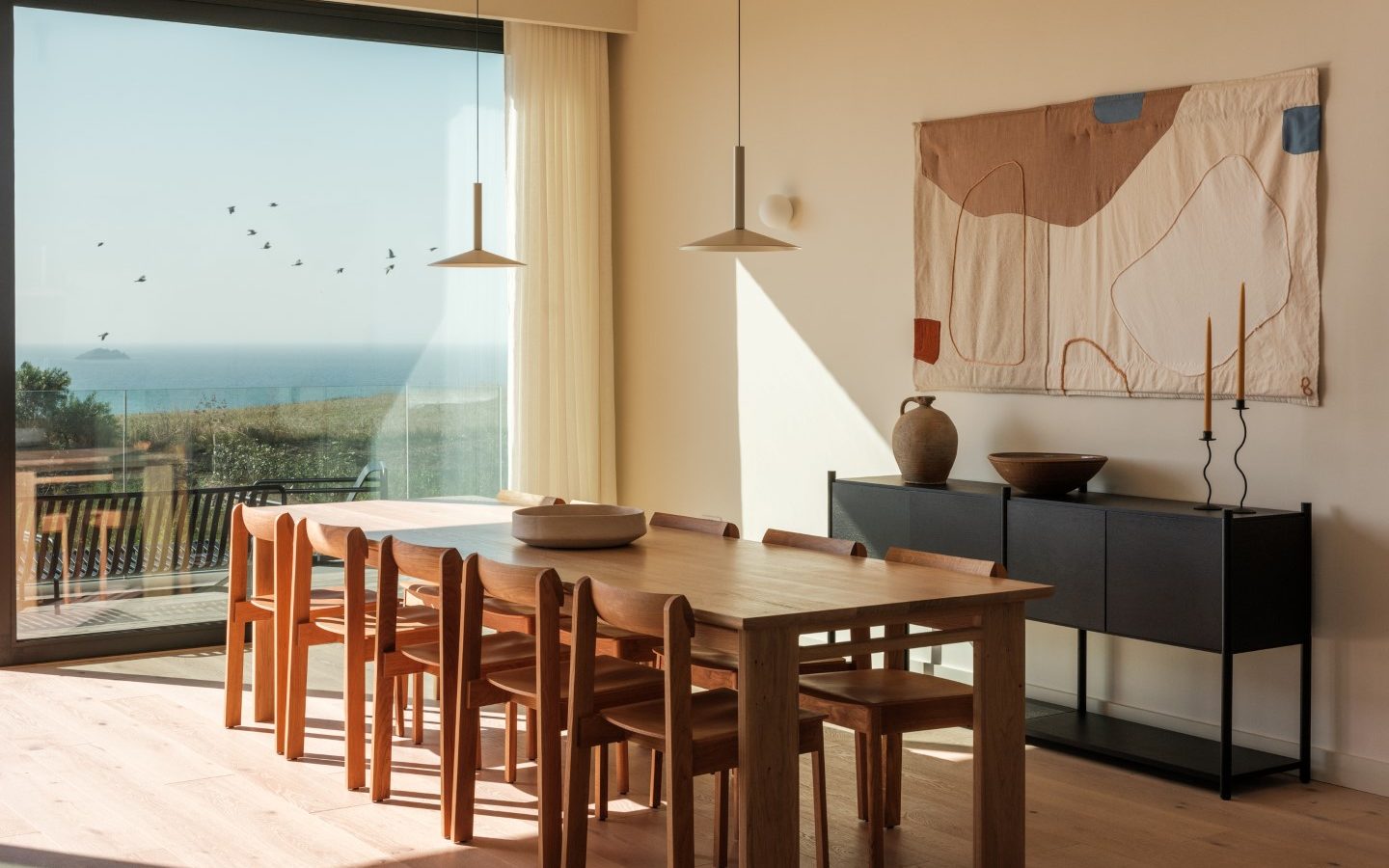 Spacious dining room with a wooden table, coastal view of Highcliffe, modern art, sideboard, and pendant lights.