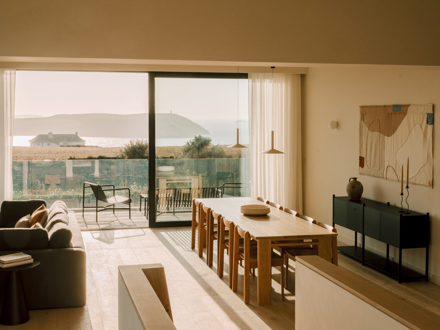 Sunlit room with a long dining table, sofa, and modern decor, overlooking a scenic view through large windows.