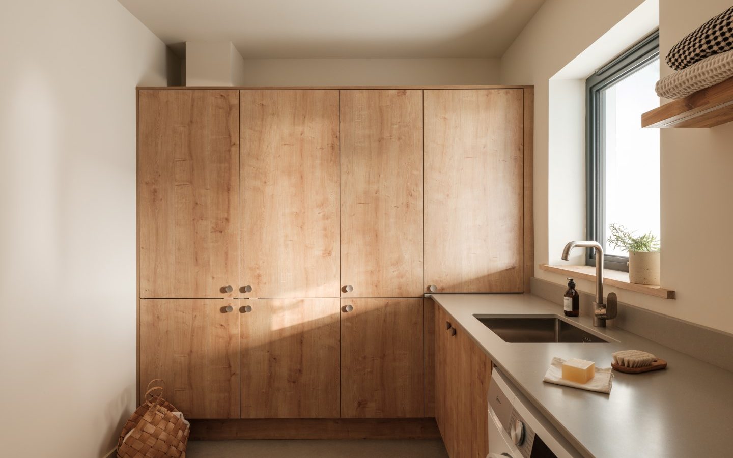 A modern Anterre laundry room with wooden cabinets, a washing machine, a sink, and a window overlooks Highcliffe.