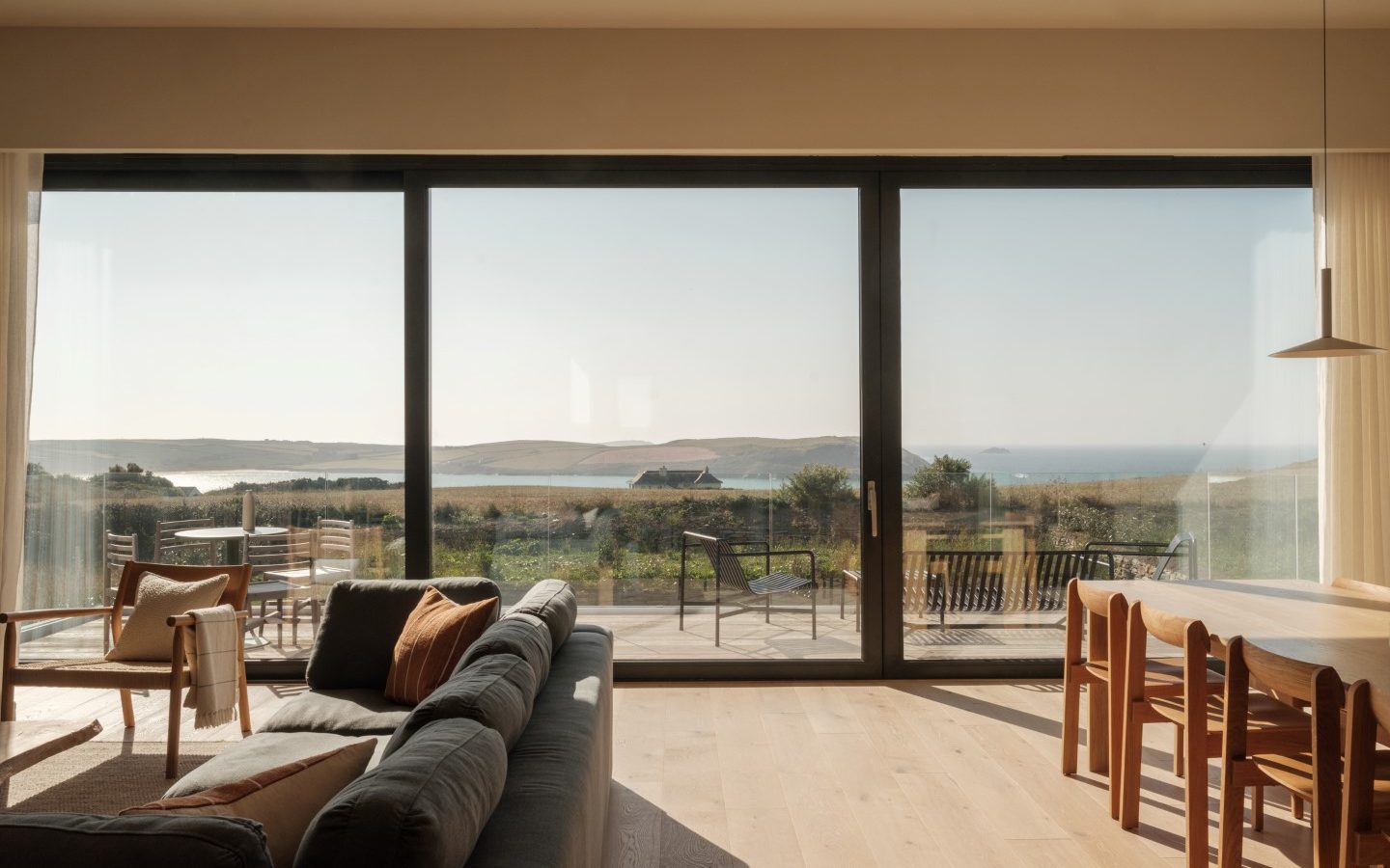 A sunlit Anterre room with a sofa and dining table overlooks Highcliffe's sea and dunes through large glass doors.