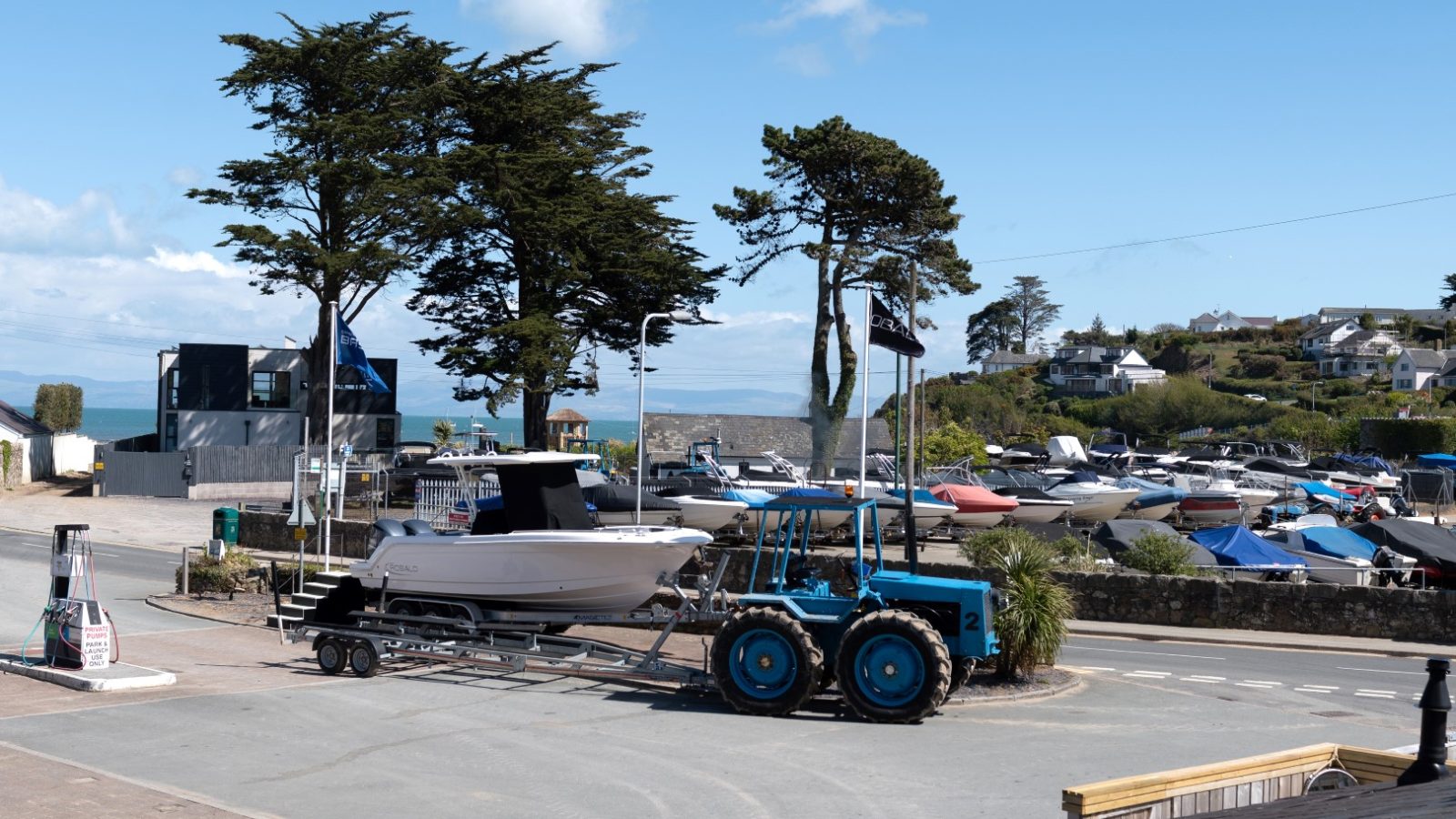 Blue tractor towing a boat on a trailer near Arfor Abersoch, with parked boats and houses against the clear blue sky.