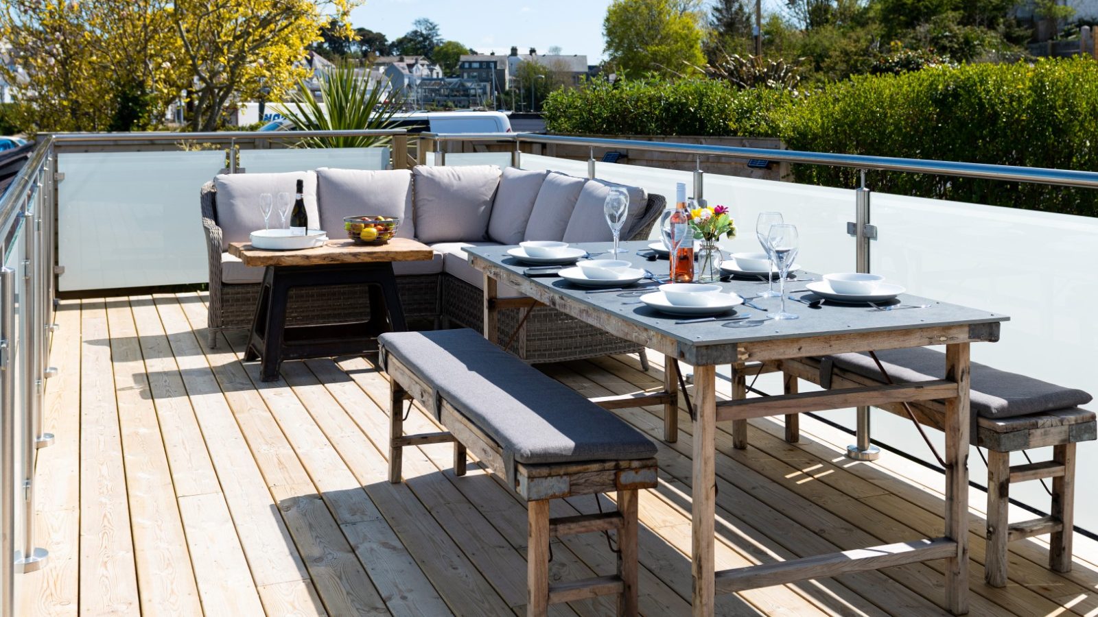 Outdoor wooden deck in Arfor Abersoch with a dining table for six, a corner sofa, bench, and a small tree in the background.