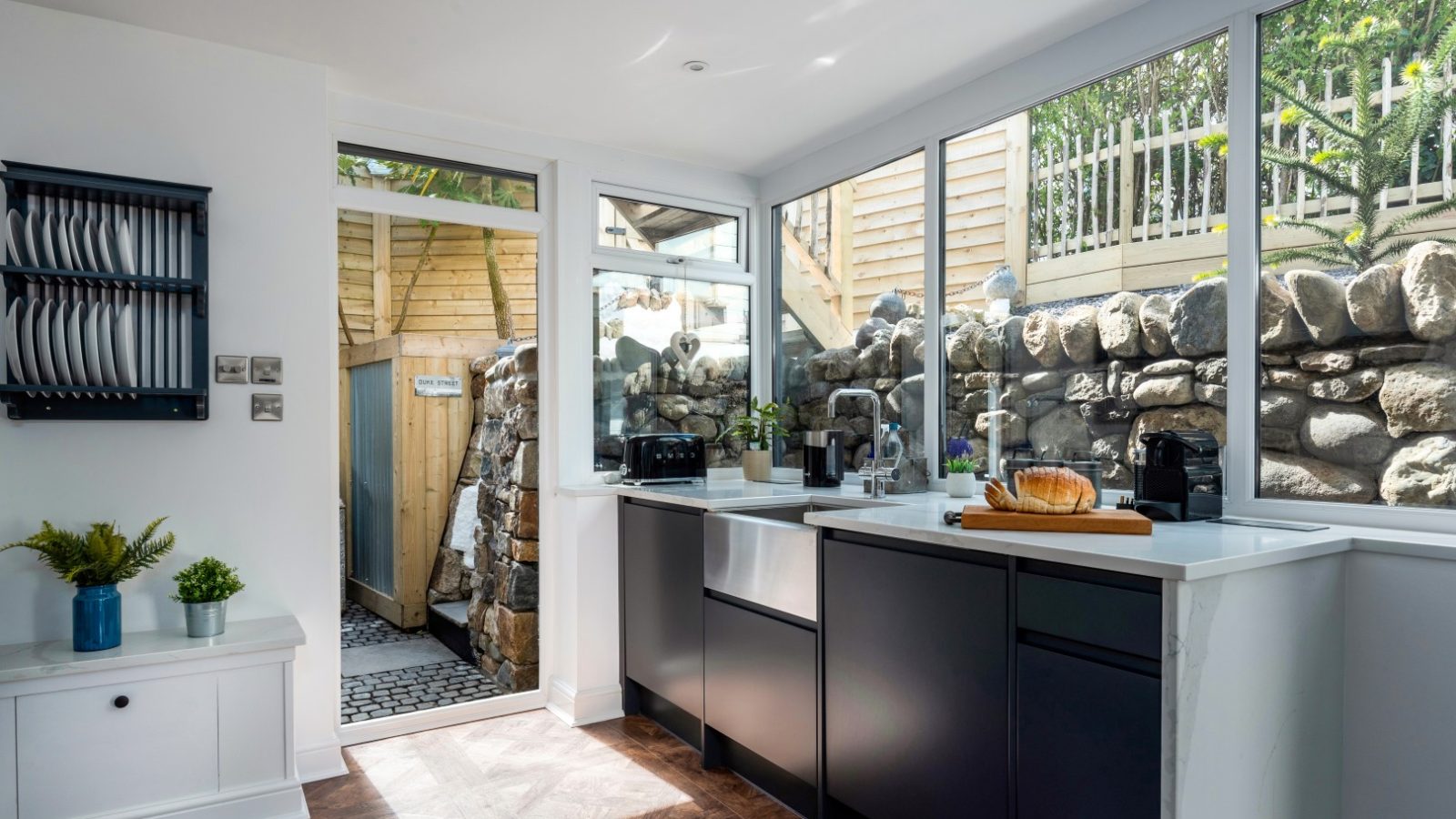 Modern kitchen with white and black cabinets, a cutting board with bread, large windows, and an Abersoch stone garden path view.