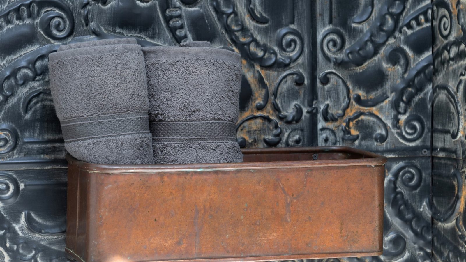 Two rolled grey towels in a brown metallic holder against a decorative dark Arfor pattern.