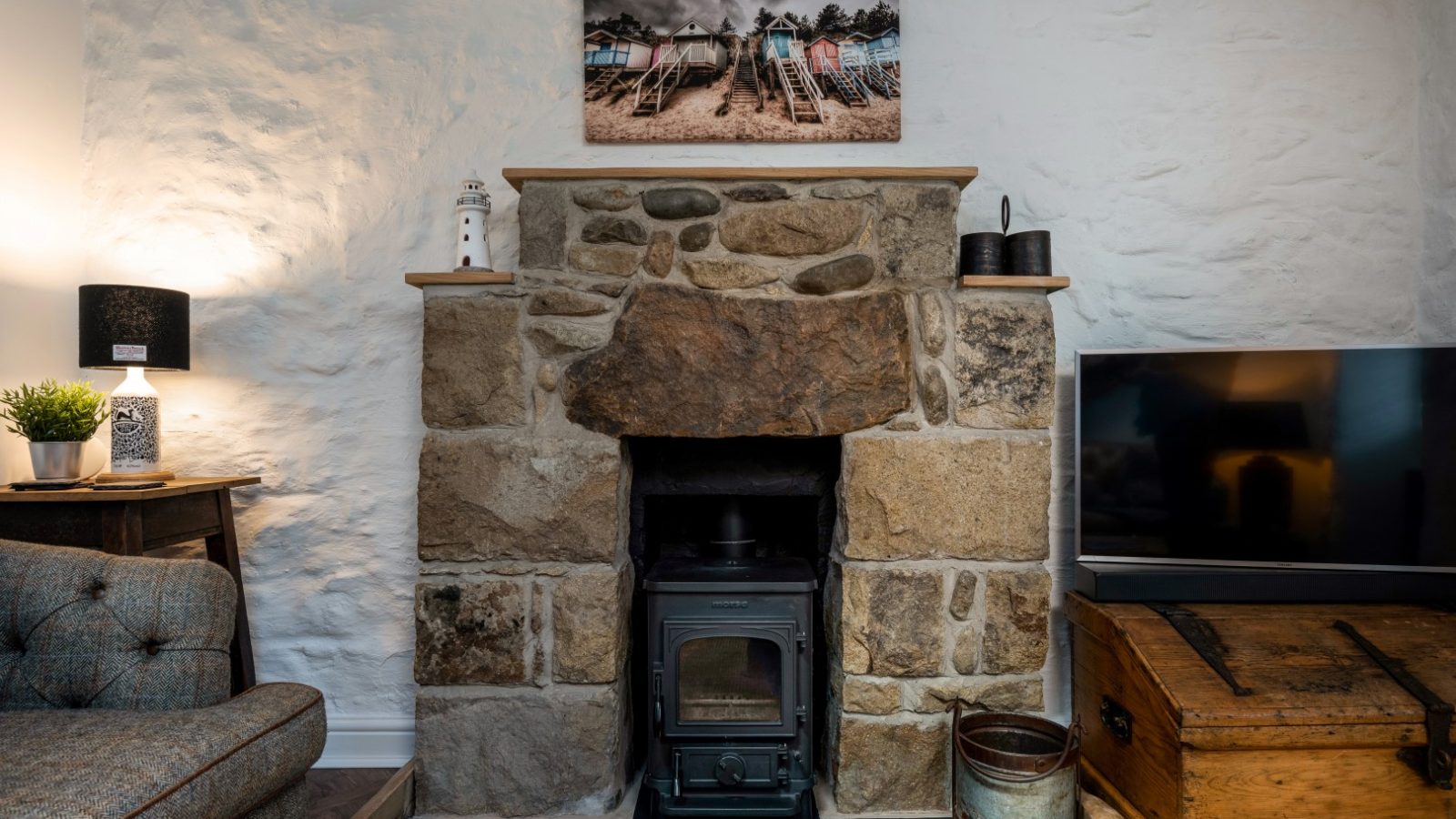 Stone fireplace with a wood-burning stove, flanked by a lamp and TV. An Arfor Abersoch landscape photo adorns the white wall.