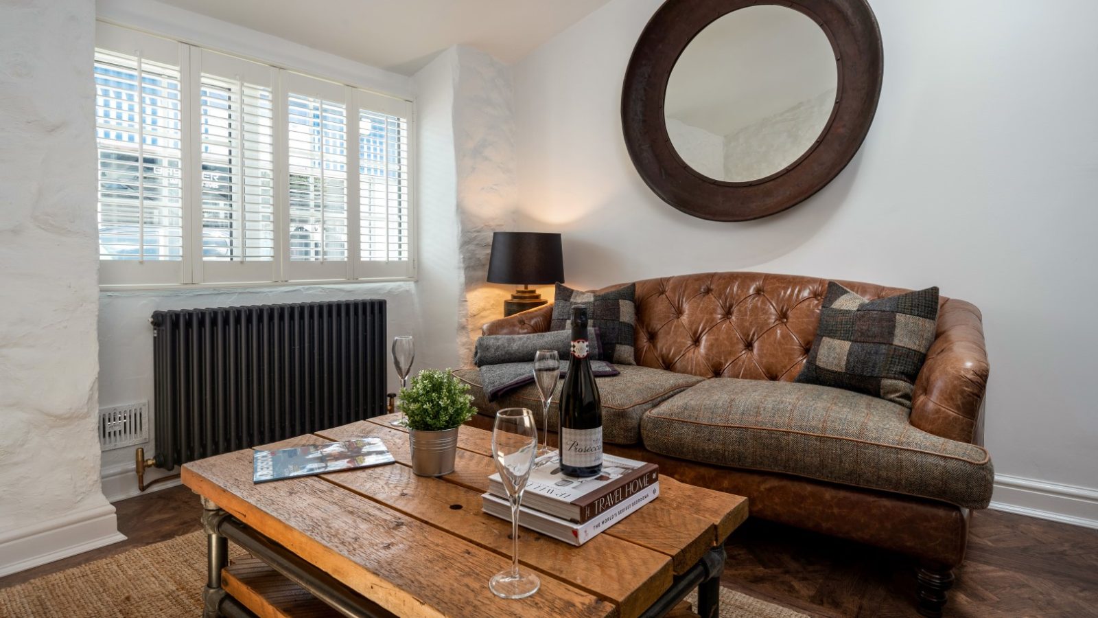 Arfor Abersoch: Cozy living room with a brown leather sofa, wooden coffee table, circular mirror, wine bottle, and lamp by the window.