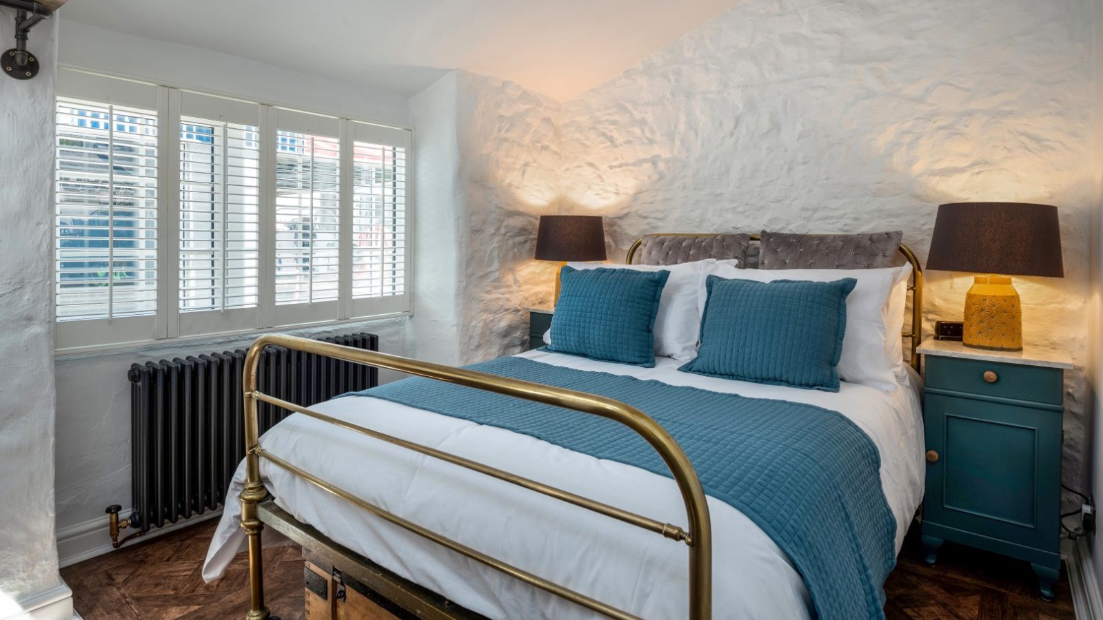 A cozy bedroom in Abersoch features a brass bed, blue bedding, two lamps, a radiator, and shuttered windows in a white-walled room.