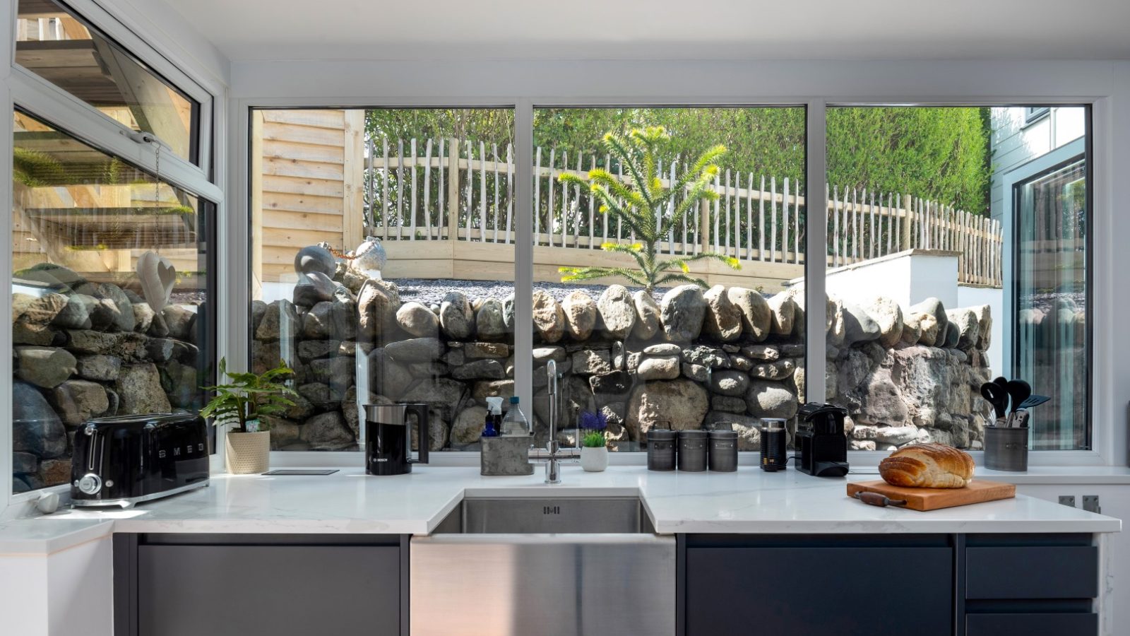 Modern kitchen with large windows, stone backsplash, stainless steel sink, and black appliances. View of Abersoch stone wall.