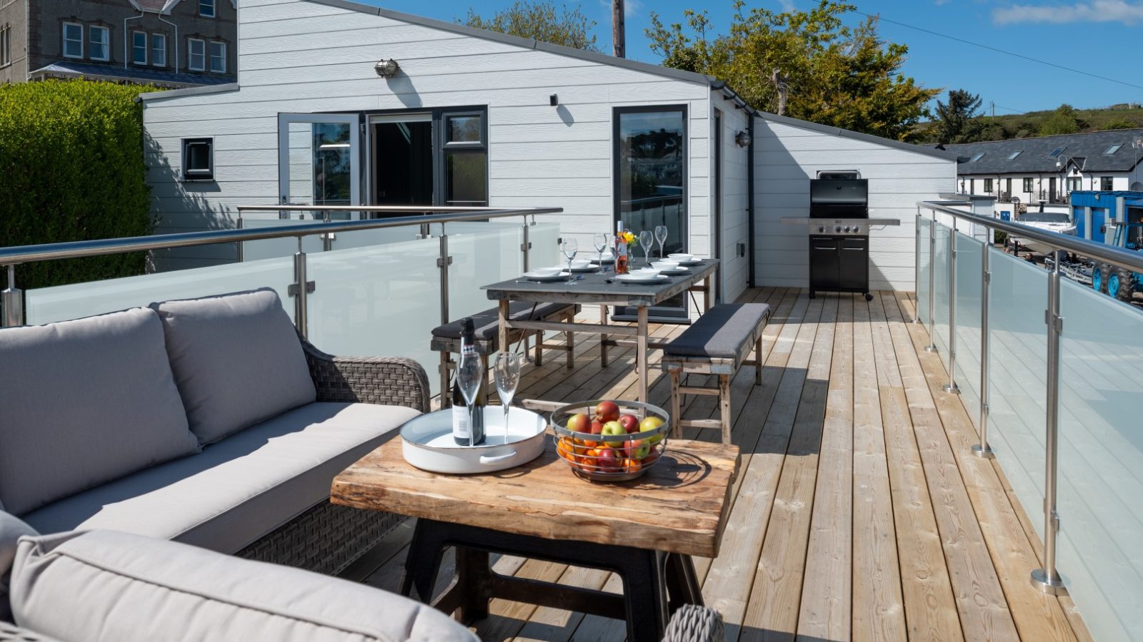 Rooftop patio in Abersoch with wicker sofa, wooden table of fruit, dining set, grill, and view of buildings and trees under a clear sky.