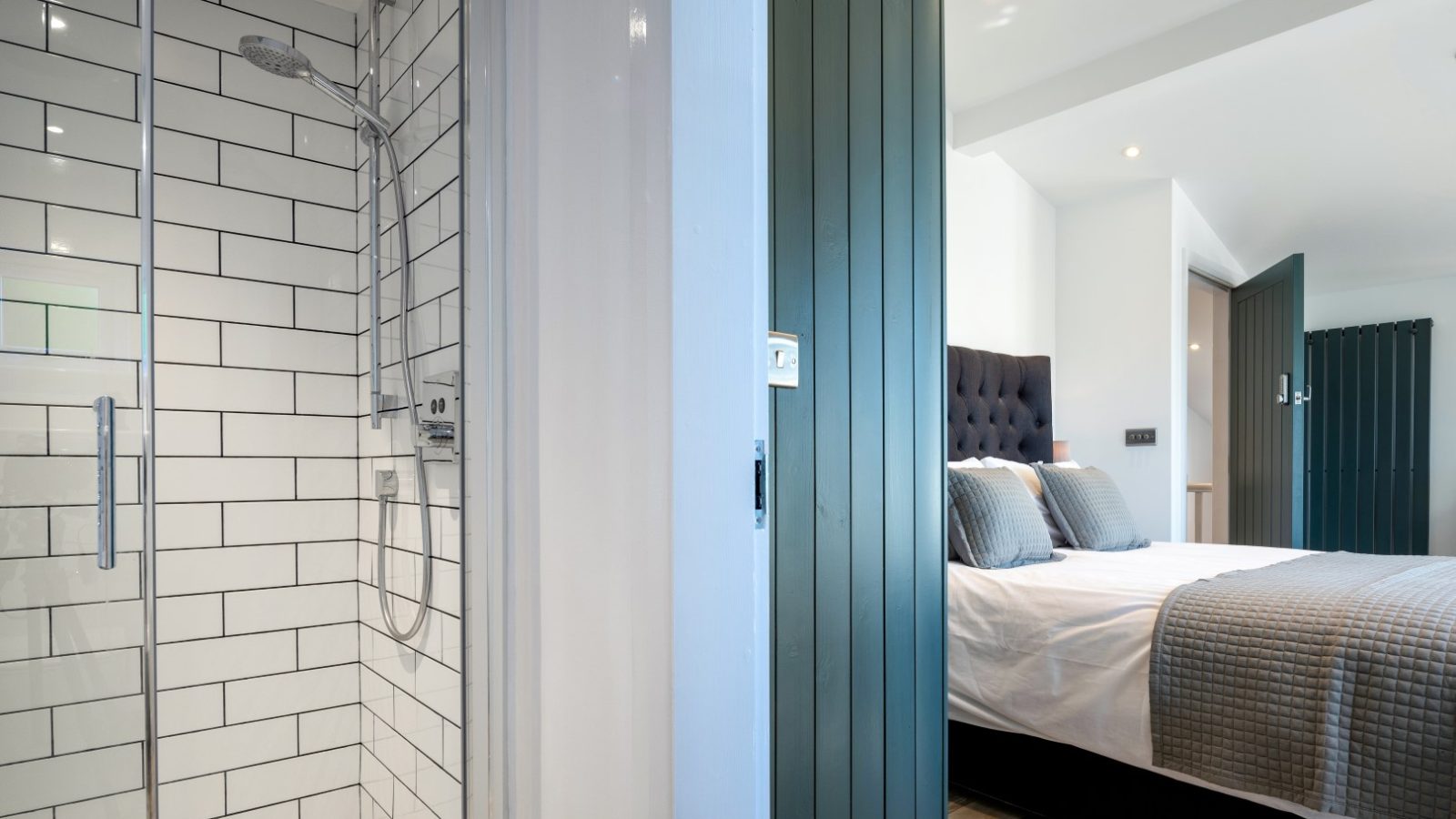 View of a bedroom with a neatly made bed and an adjacent shower area in Arfor, featuring white subway tiles and glass door.