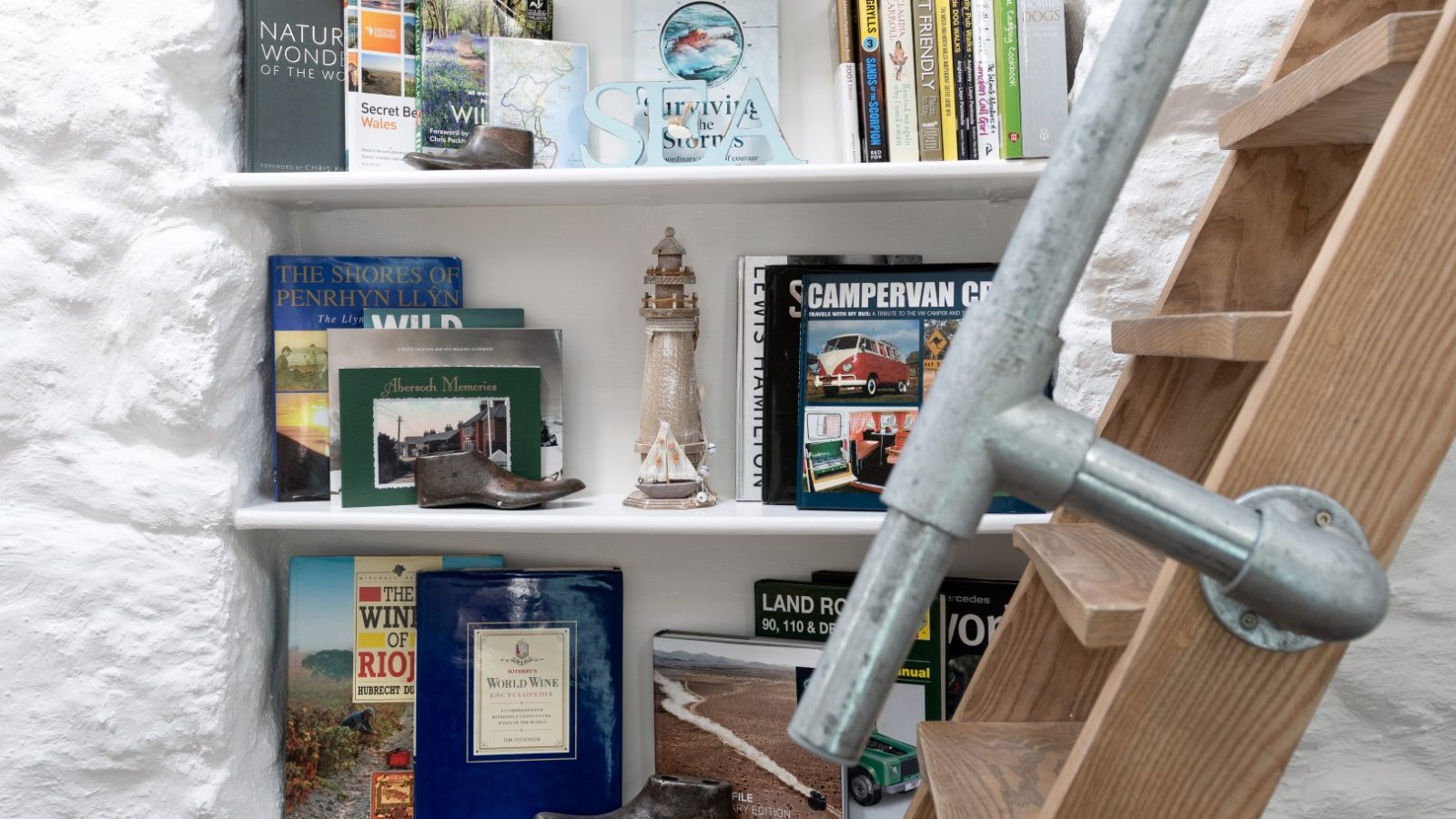 Bookshelf with travel and wine books, Arfor Abersoch model lighthouse, wooden shoe, and rustic ladder by a white stone wall.