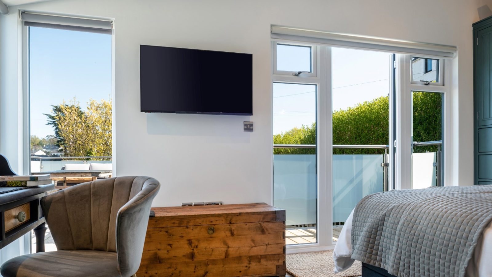 Modern bedroom in Abersoch with a wall-mounted TV, large windows, desk, wooden chest, and bed by the balcony.