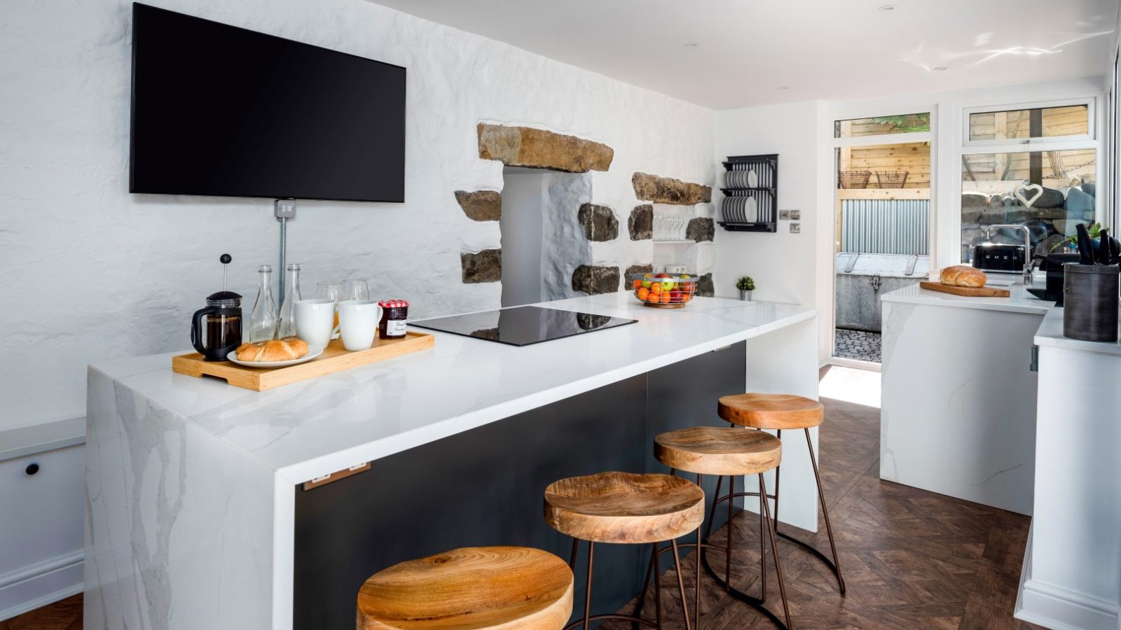 Modern kitchen in Abersoch with white walls, a black-mounted TV, wooden stools, and a central island with cooktop and fruit bowl.