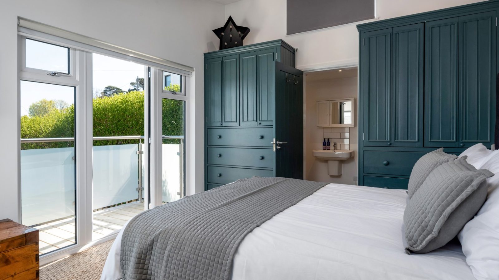 A bedroom with a double bed, green wardrobes, and an ensuite. Glass doors open to a balcony overlooking Abersoch’s greenery.