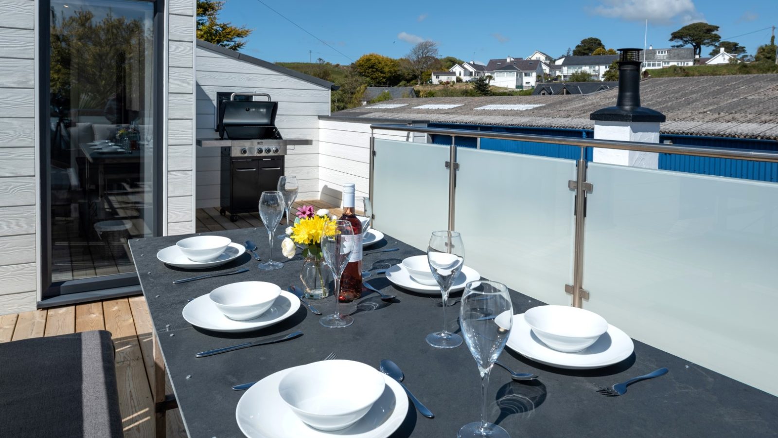 Outdoor dining setup on a patio with a table, white plates, glasses, cutlery, flowers, wine, and an Arfor barbecue grill.