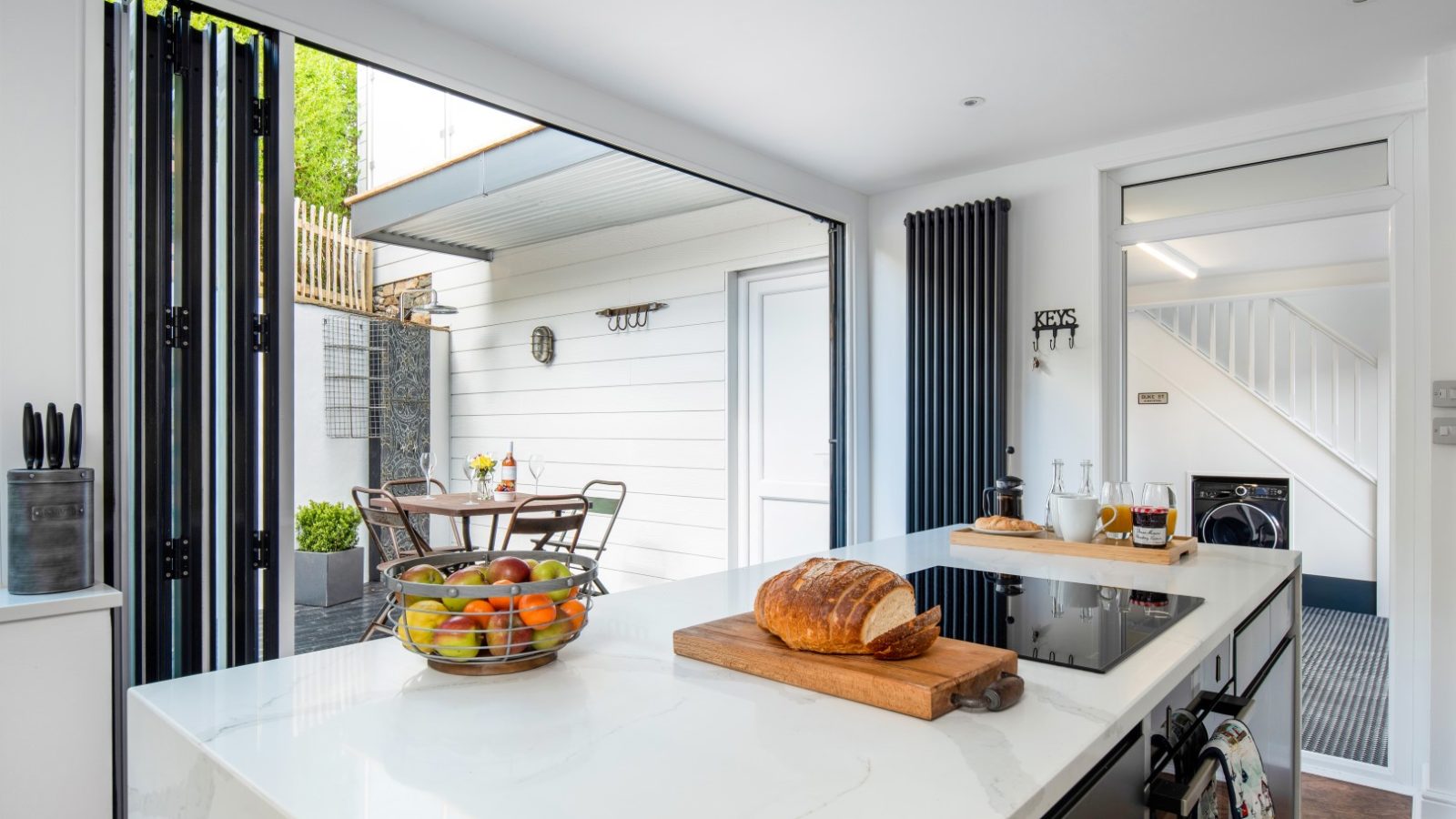 Modern kitchen in Abersoch with marble island, induction cooktop, bread on a board, and a bowl of fruit opening to patio dining.