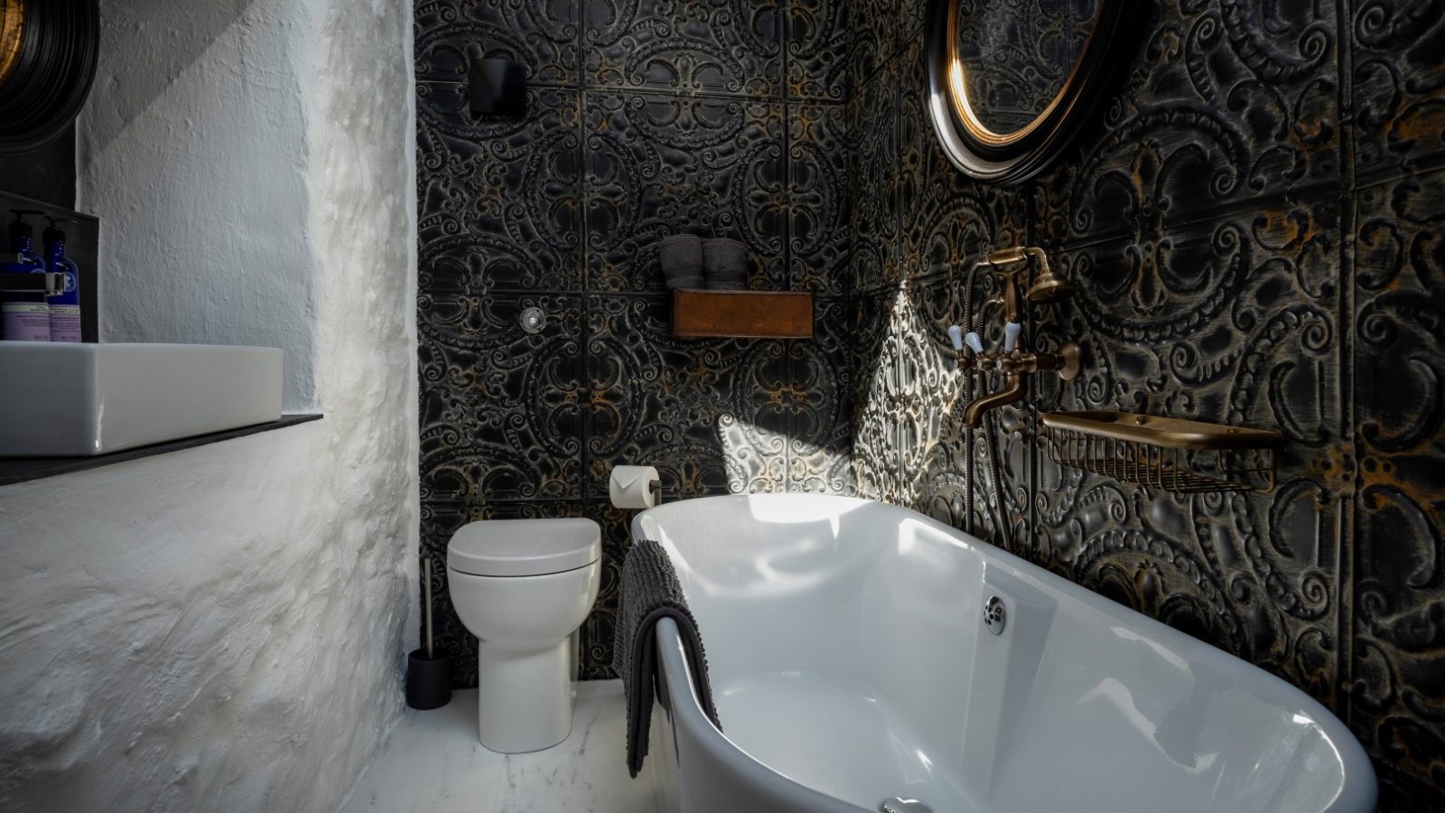 Bathroom in Abersoch with a white freestanding tub, round mirror, ornate dark tiles, white toilet, and small sink.