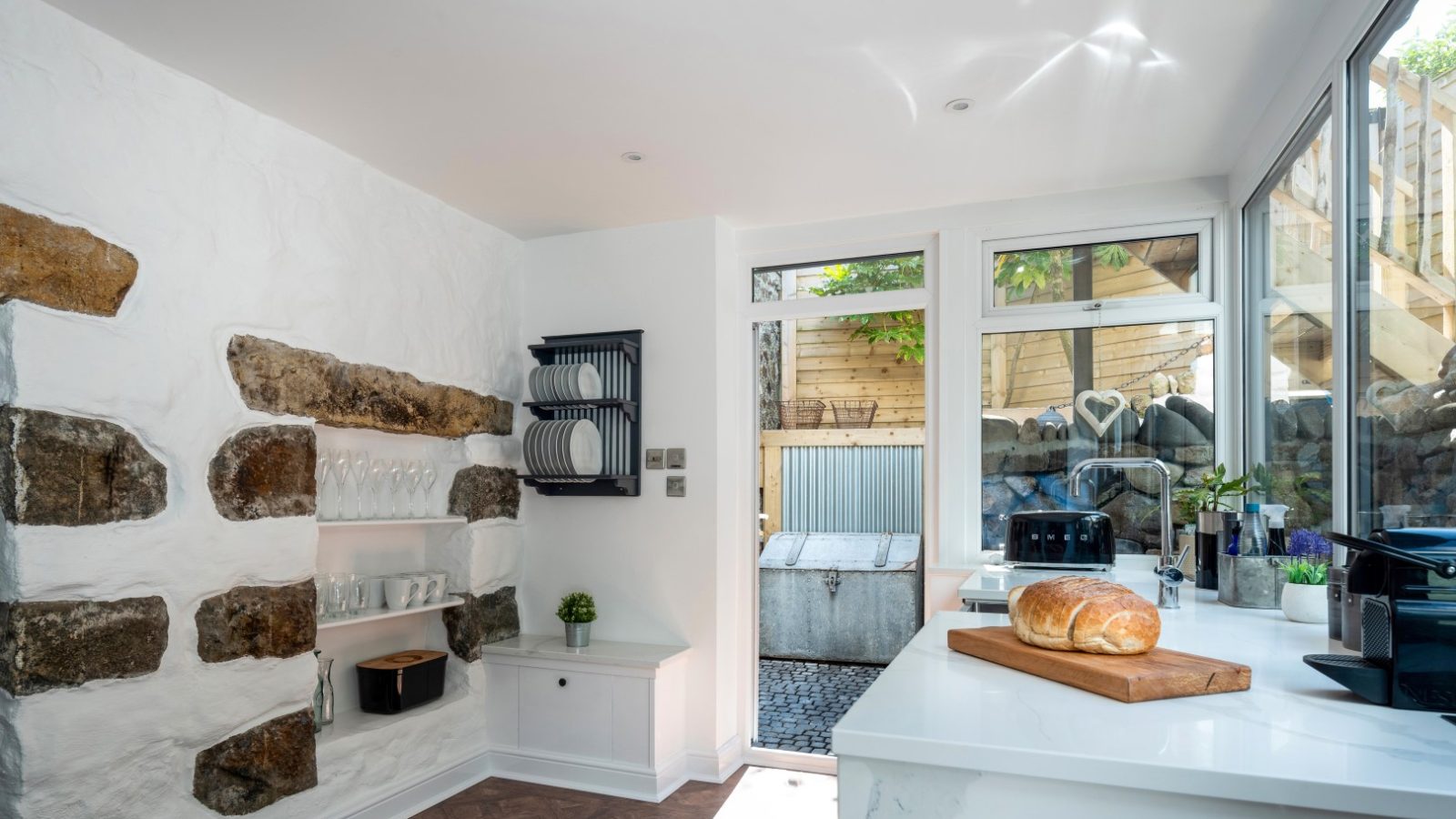 Bright kitchen with rustic stone wall, Abersoch charm, shelves with dishes, countertop with bread, large windows, and outdoor door.