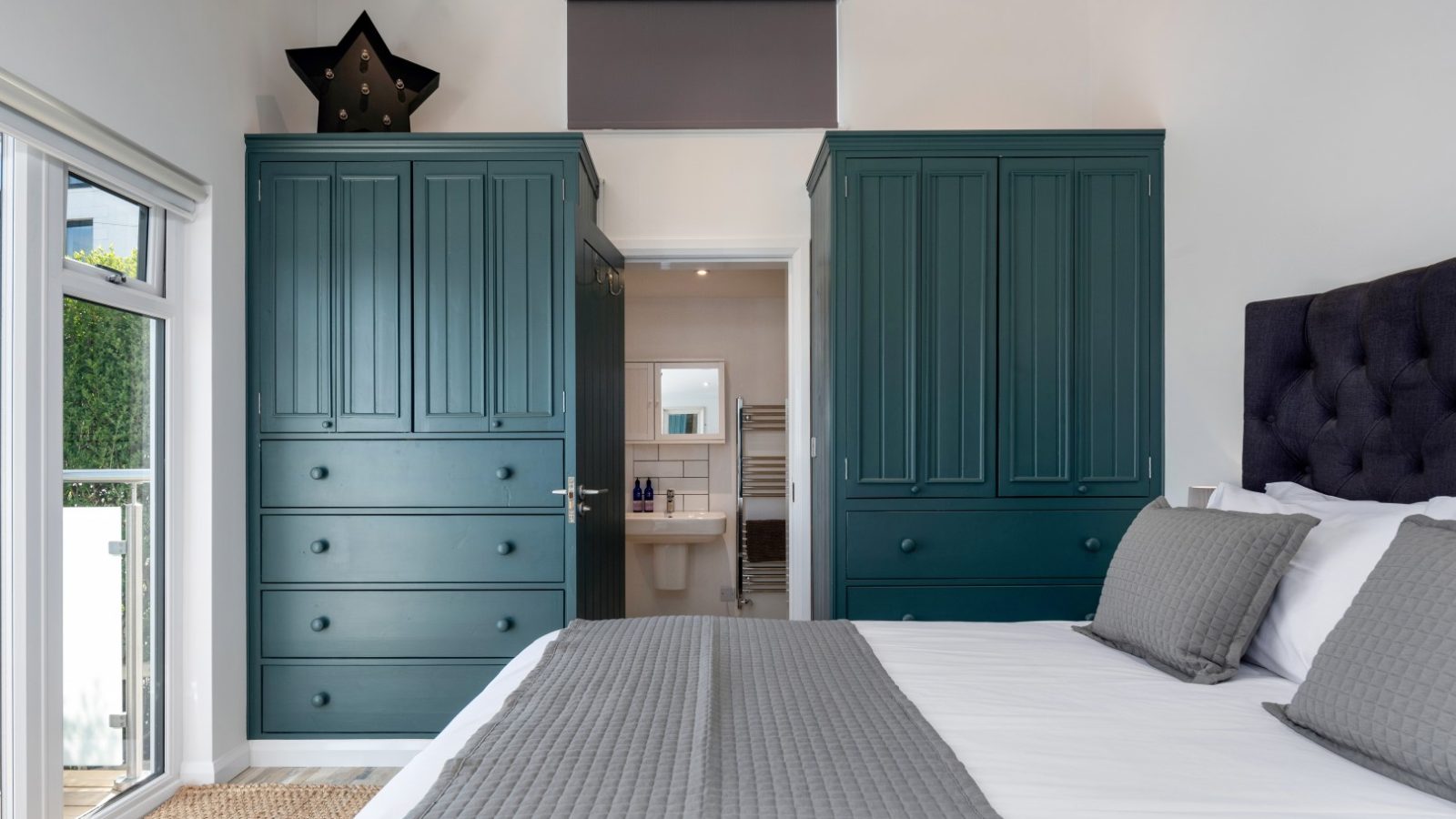 Bedroom with a neatly made bed, gray bedding, teal wardrobes, and a glimpse of the Arfor Abersoch bathroom through an open door.