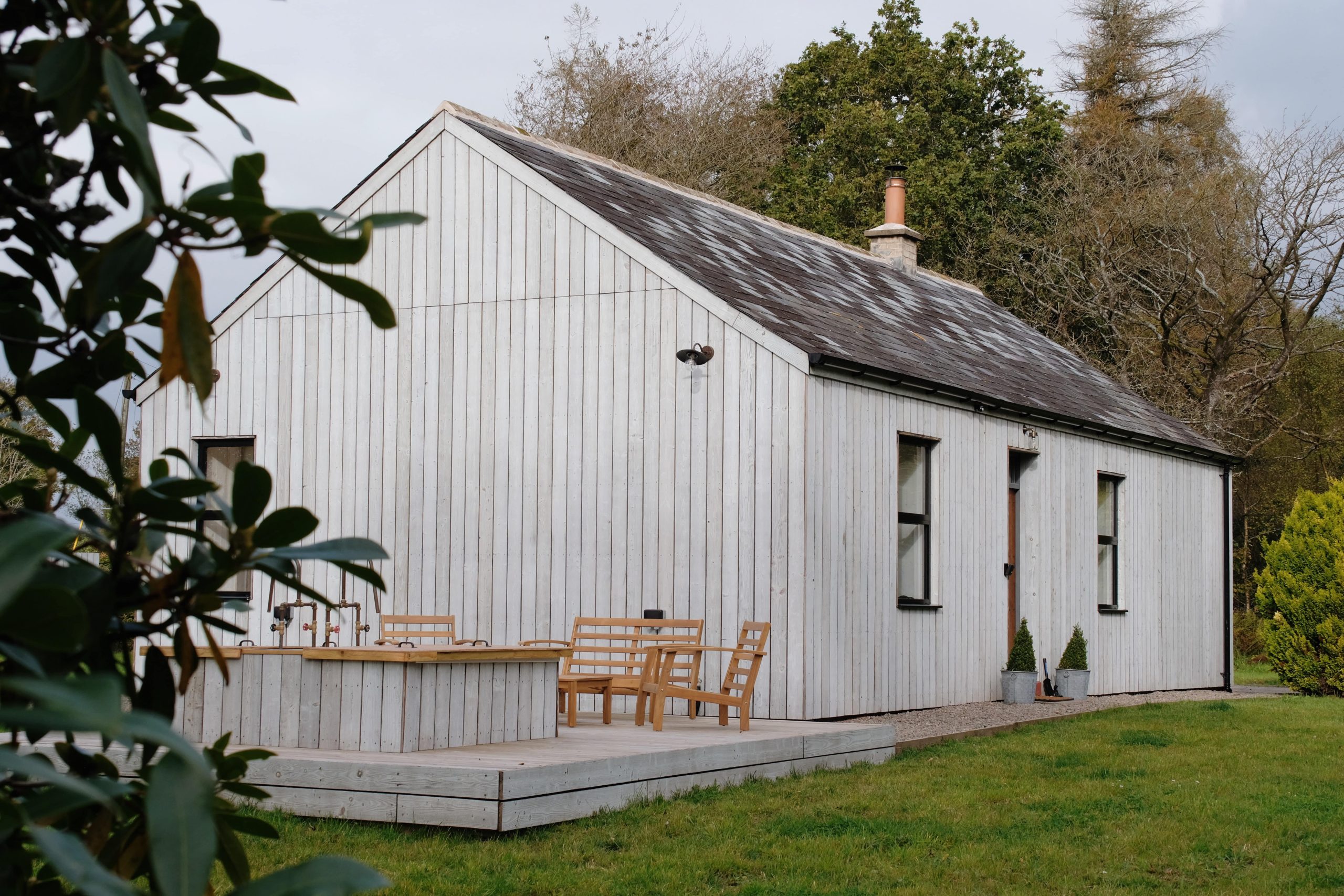 Charming white wooden house with a patio for Digital Detox Stays, nestled in greenery under an overcast sky.