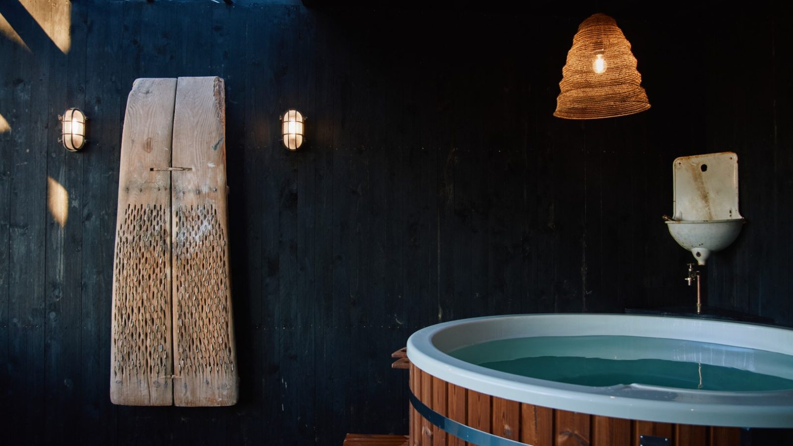 Rustic spa setting at Bonington Farm with a round wooden hot tub, woven lampshade, and textured wall decor against dark wood walls.