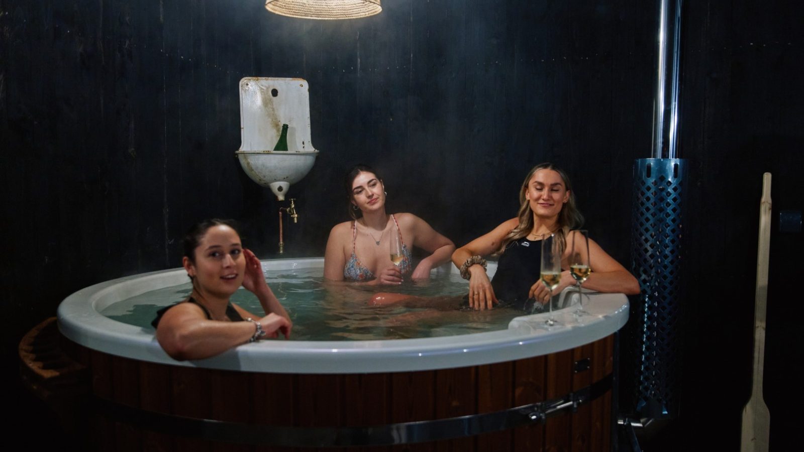 Three women unwind in a steaming wooden hot tub at Bonington Farm, with plants overhead and a paddle beside them.