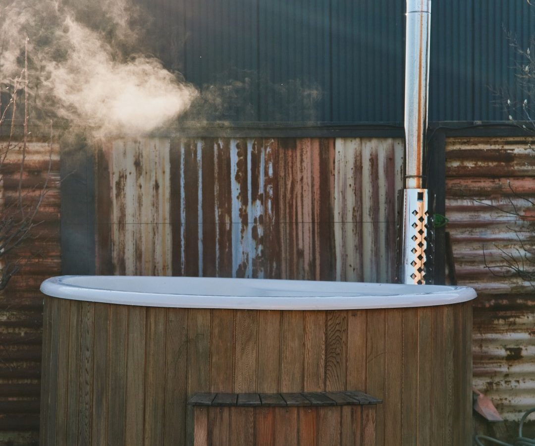 Wooden hot tub with steps outside, emitting steam at Bonington Farm. Background: wooden fence and a metal-roofed building.