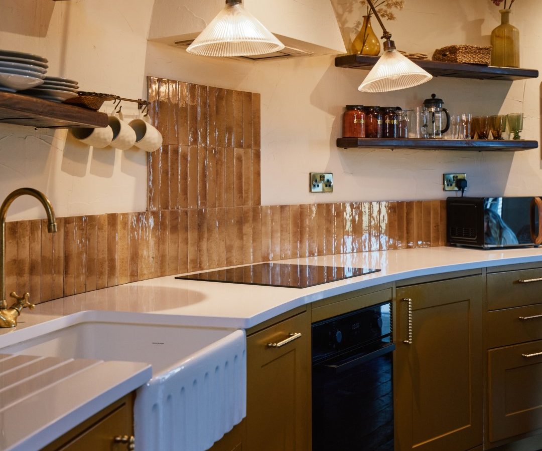 A cozy kitchen with mustard cabinets, open shelves, a Bonington Farm sink, and a brown tiled backsplash.