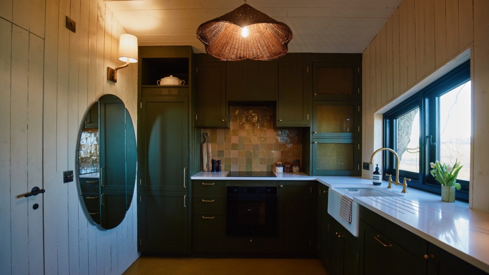 Cozy Bonington Farm kitchen with dark green cabinets, round mirror, woven light fixture, and a large window with tiled backsplash.