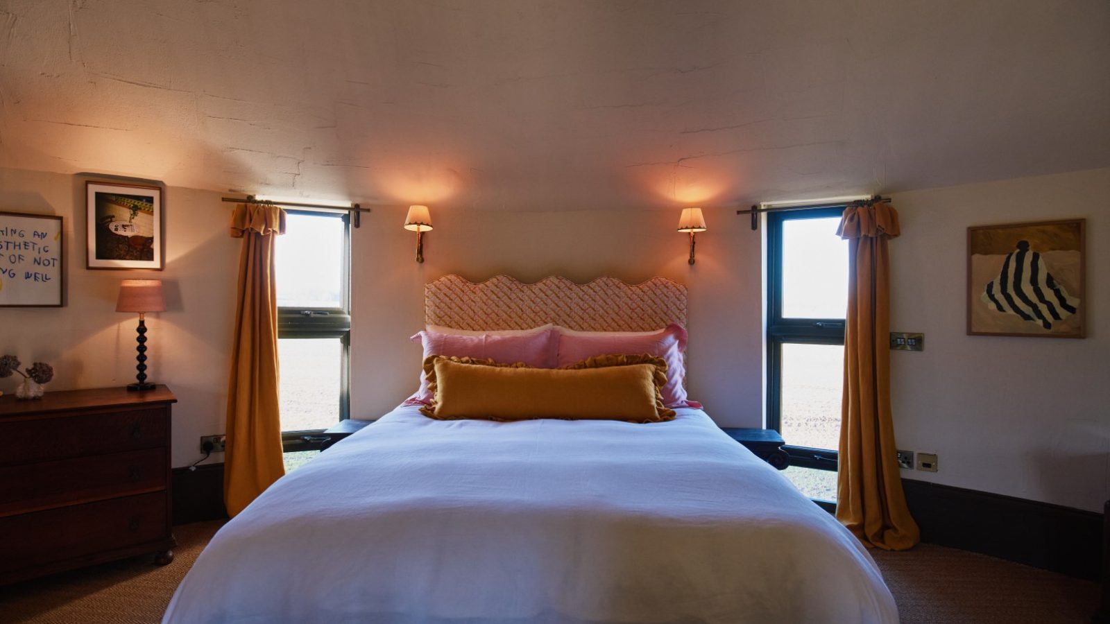 A cozy Bonington Farm bedroom with a double bed, white bedding, yellow accents, two windows, lamps, and framed artwork.