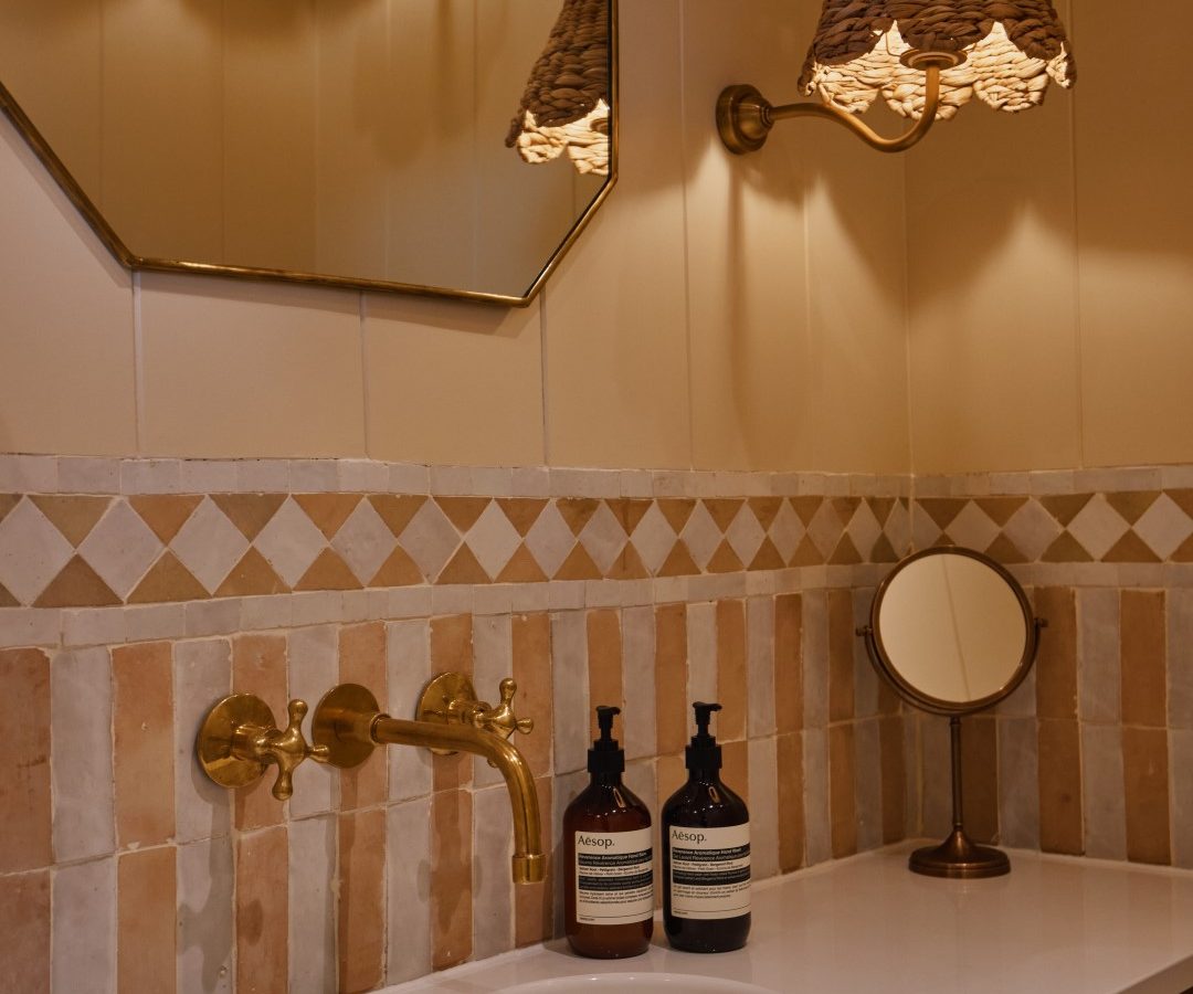 Elegant Bonington Farm bathroom with brass fixtures, a large mirror, woven wall sconce, and decorative tiles.