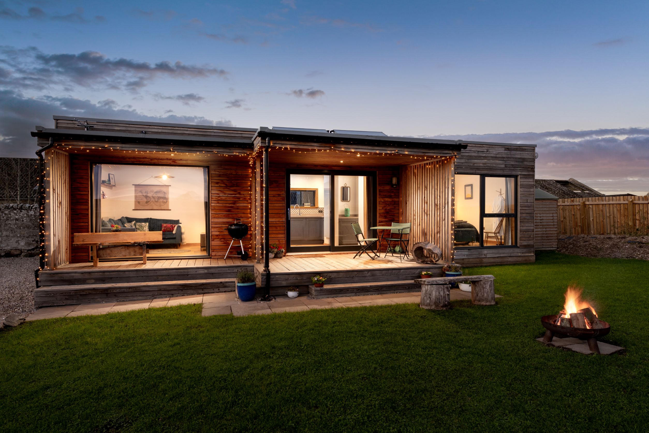 Modern cabin exterior with large windows, string lights, and a wooden deck offers a perfect Digital Detox Stay by the fire pit.