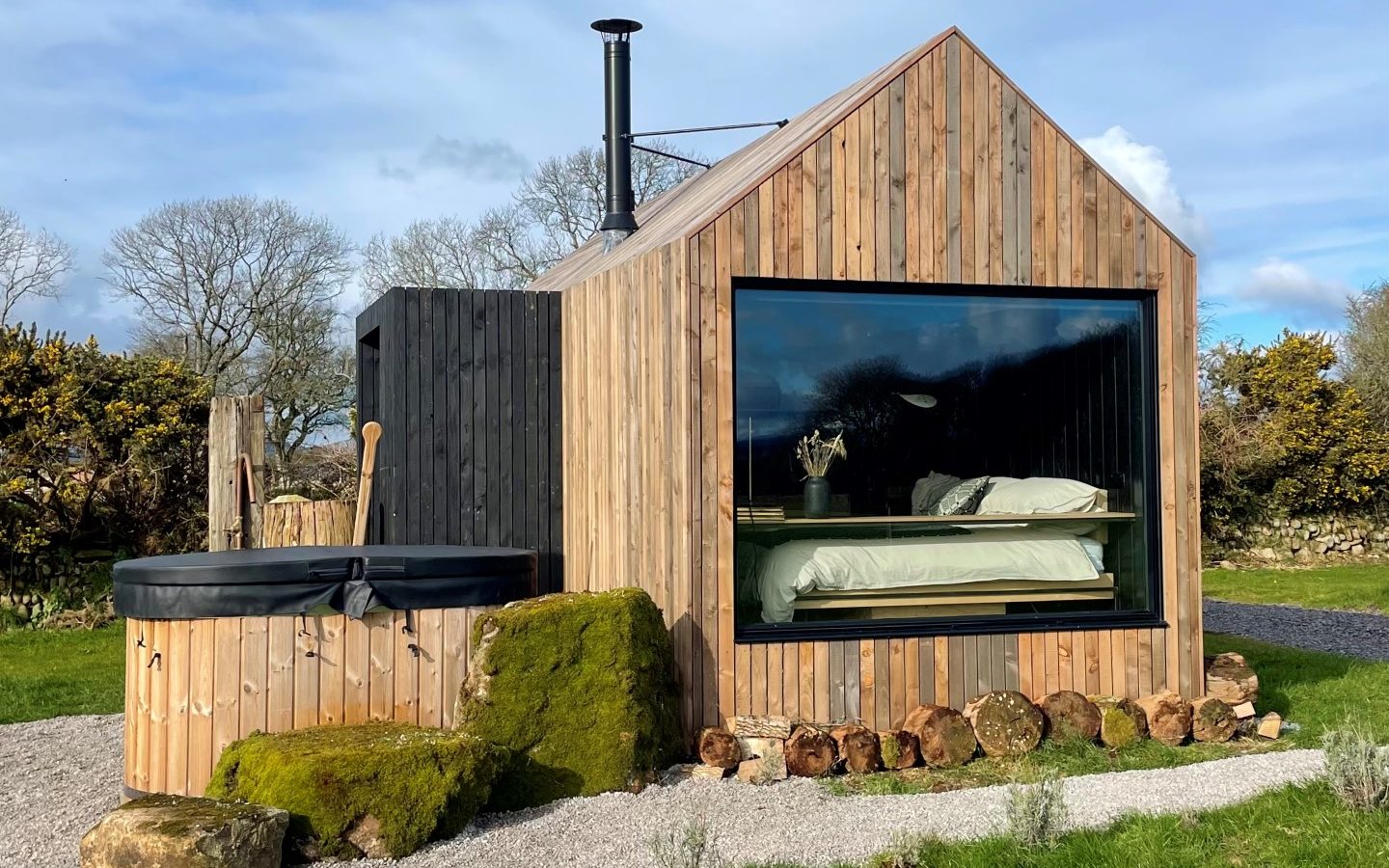 Wooden cabin with a large window showcasing a bed inside, next to an outdoor hot tub and surrounded by greenery.