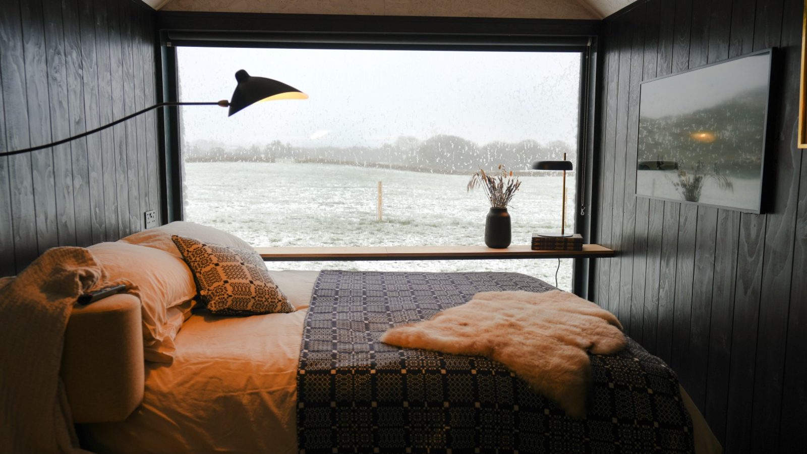 Cozy bedroom with a large window view of a snowy landscape. A bed with patterned bedding, a lamp, and a vase decorate the space.