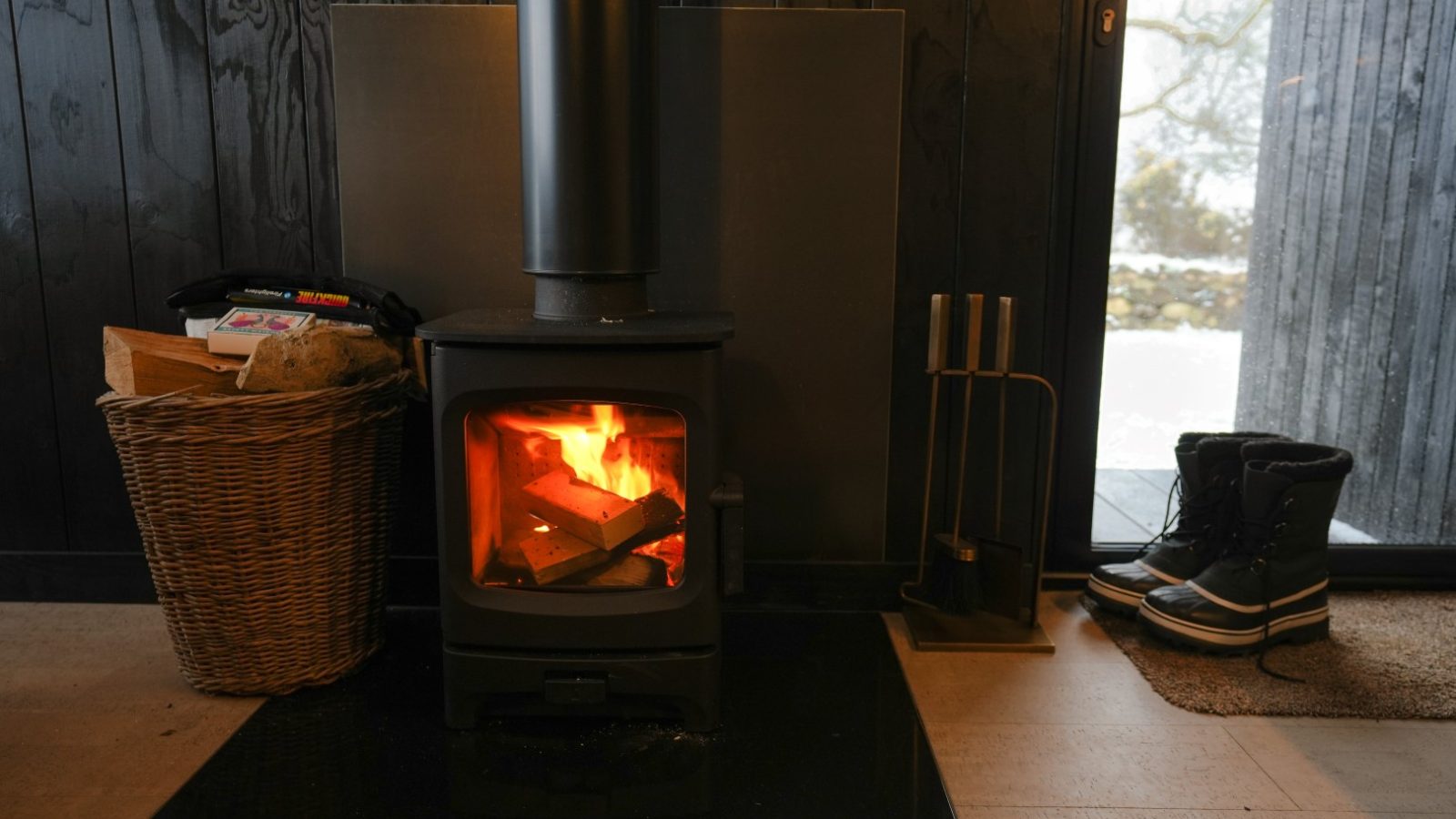 A lit wood-burning stove with a basket of logs beside it. Snow boots are placed near a glass door showing a snowy outdoors.