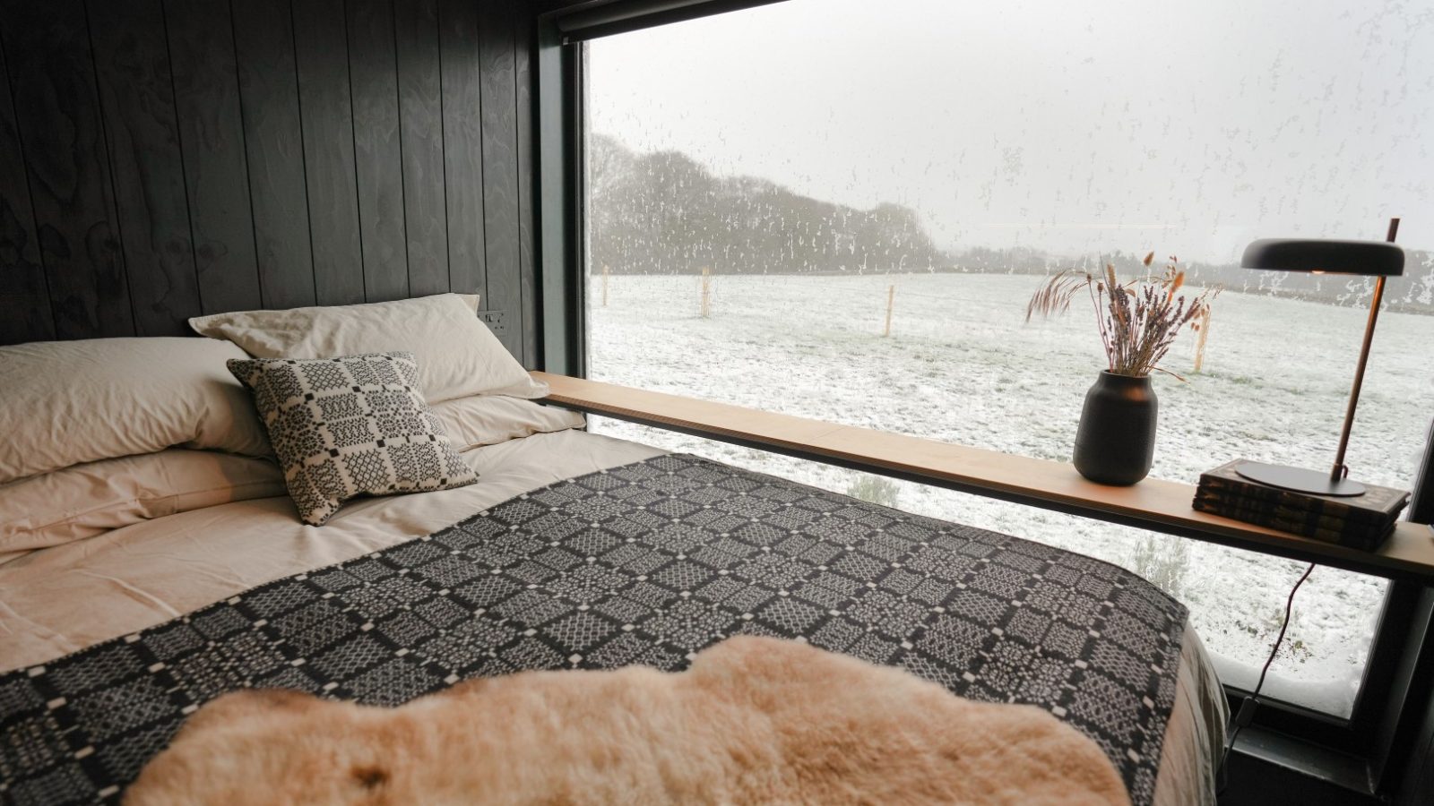 Cozy bedroom with large window view of a snowy landscape. Bed has patterned blanket, decorative pillows, and a lamp on the sill.