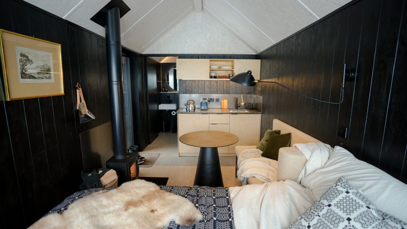 Cozy cabin interior with dark wood, small kitchen area, bed with patterned blankets, and wood-burning stove in the corner.