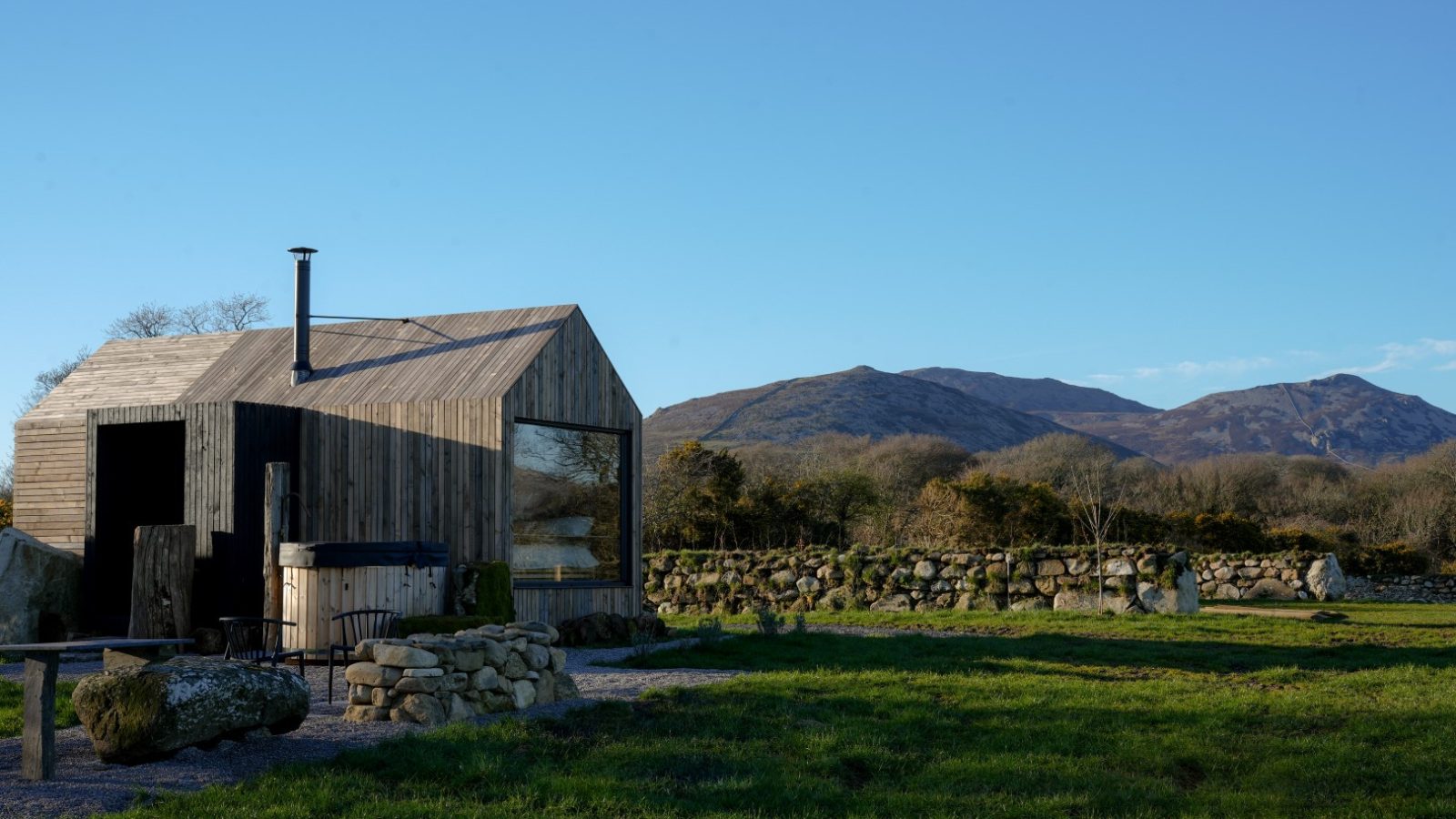 Small wooden cabin with a glass window in a grassy field, surrounded by stone walls, and mountains in the background under clear sky.