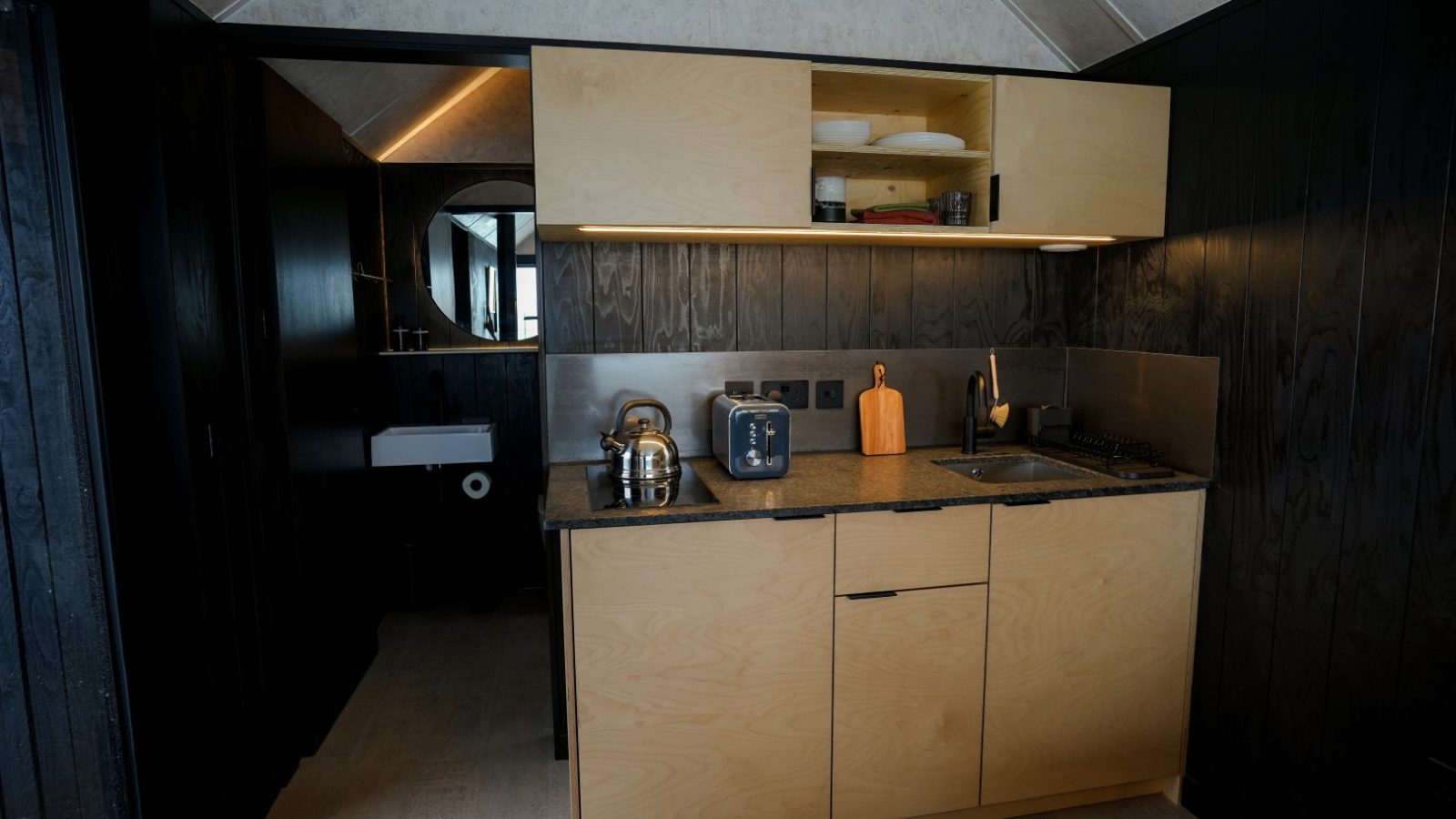 Compact kitchen with wooden cabinets, a small sink, kettle, toaster, and open shelves. Adjacent to a dark-colored wall.