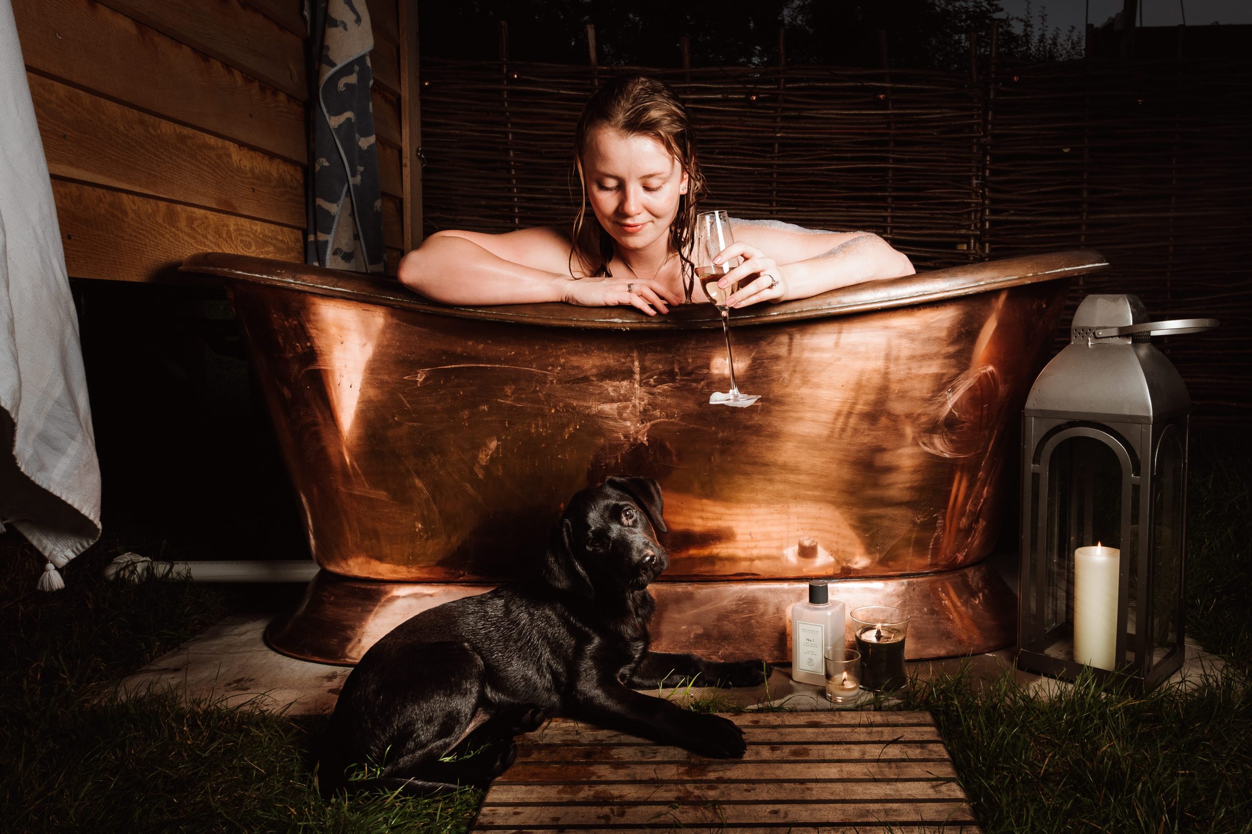 Woman enjoying a romantic outdoor soak with wine, her black dog lounging nearby, a candle flickering softly on the right.