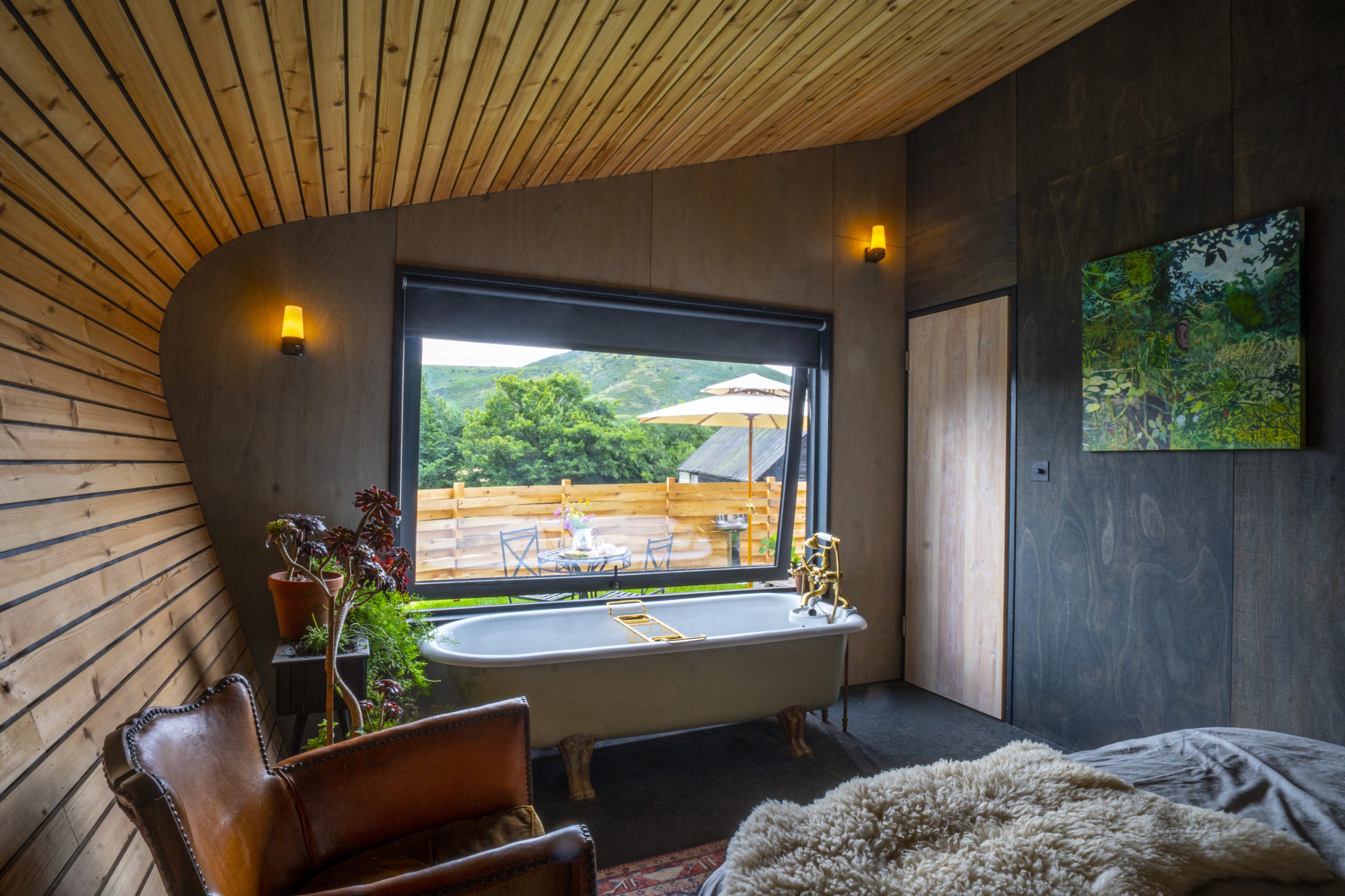 A cozy, romantic room with a standalone bathtub, wooden walls, a leather chair, and a large window overlooking a patio.