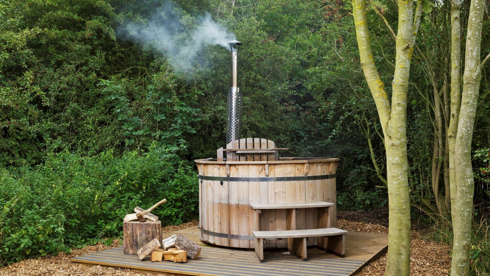 Secret Meadows: A wood-fired hot tub nestles among trees, with smoke curling from the chimney and logs stacked beside it.